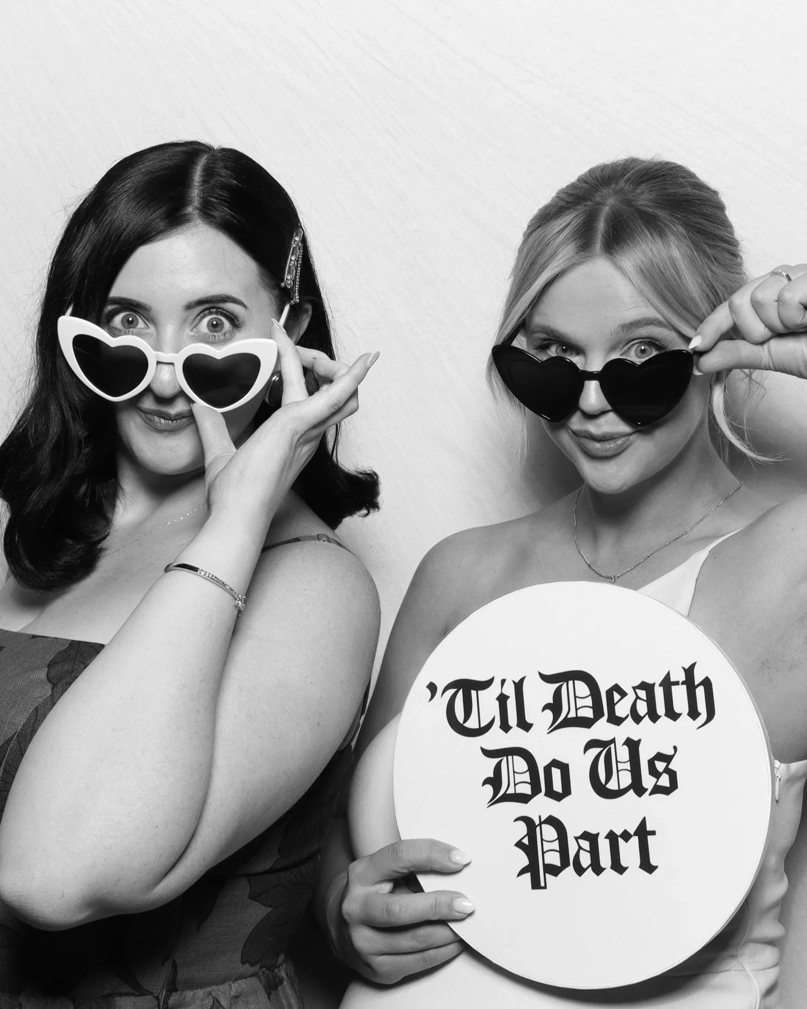Two women with styled hair and makeup wearing sunglasses, with one woman holding a sign that reads 'Till Death Do Us Part', both standing against a plain backdrop.