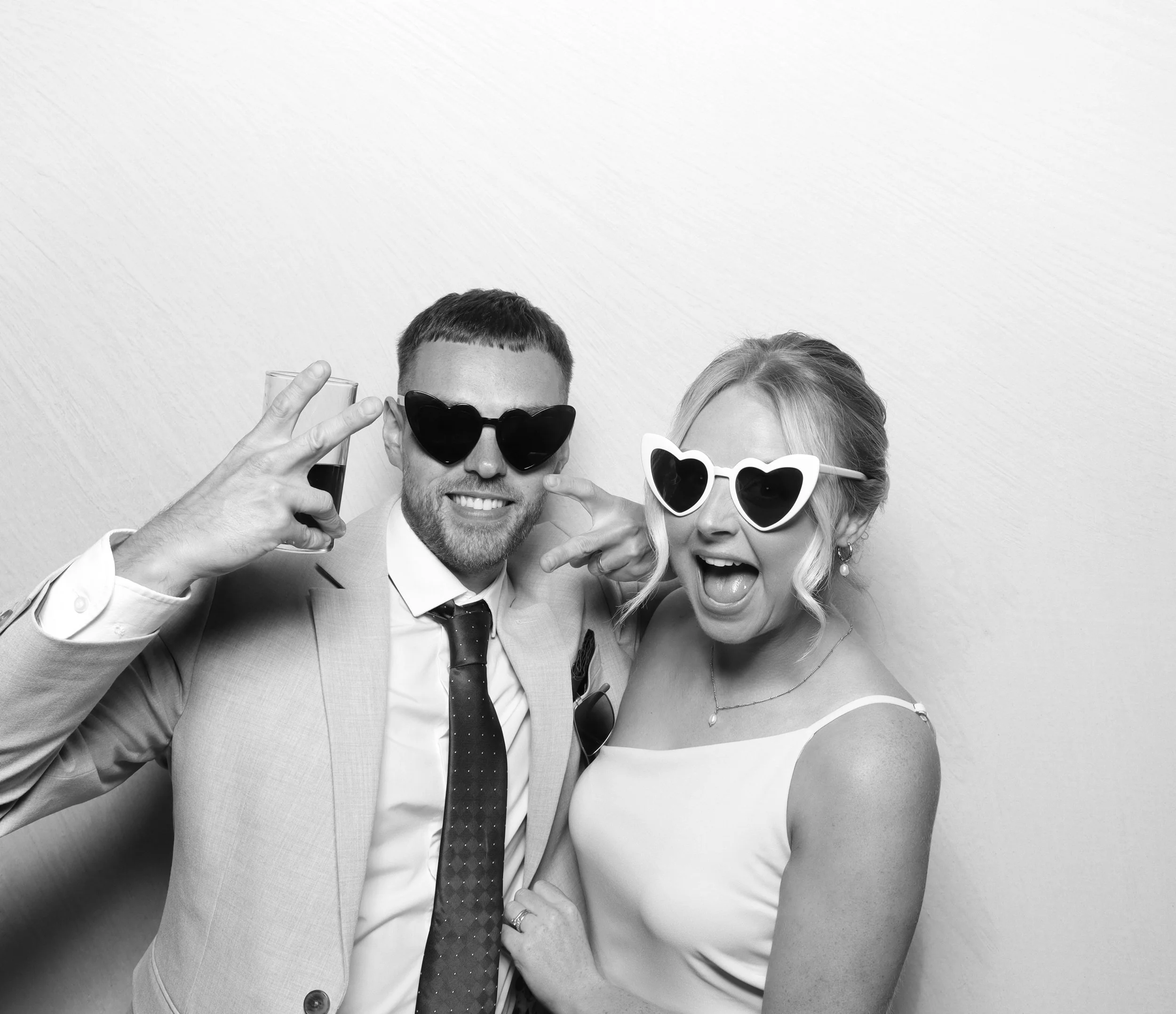 A black-and-white photo of a happy couple dressed in formal attire, wearing heart-shaped sunglasses, smiling and posing. The man holds a glass and makes a peace sign, while the woman sticks out her tongue playfully.