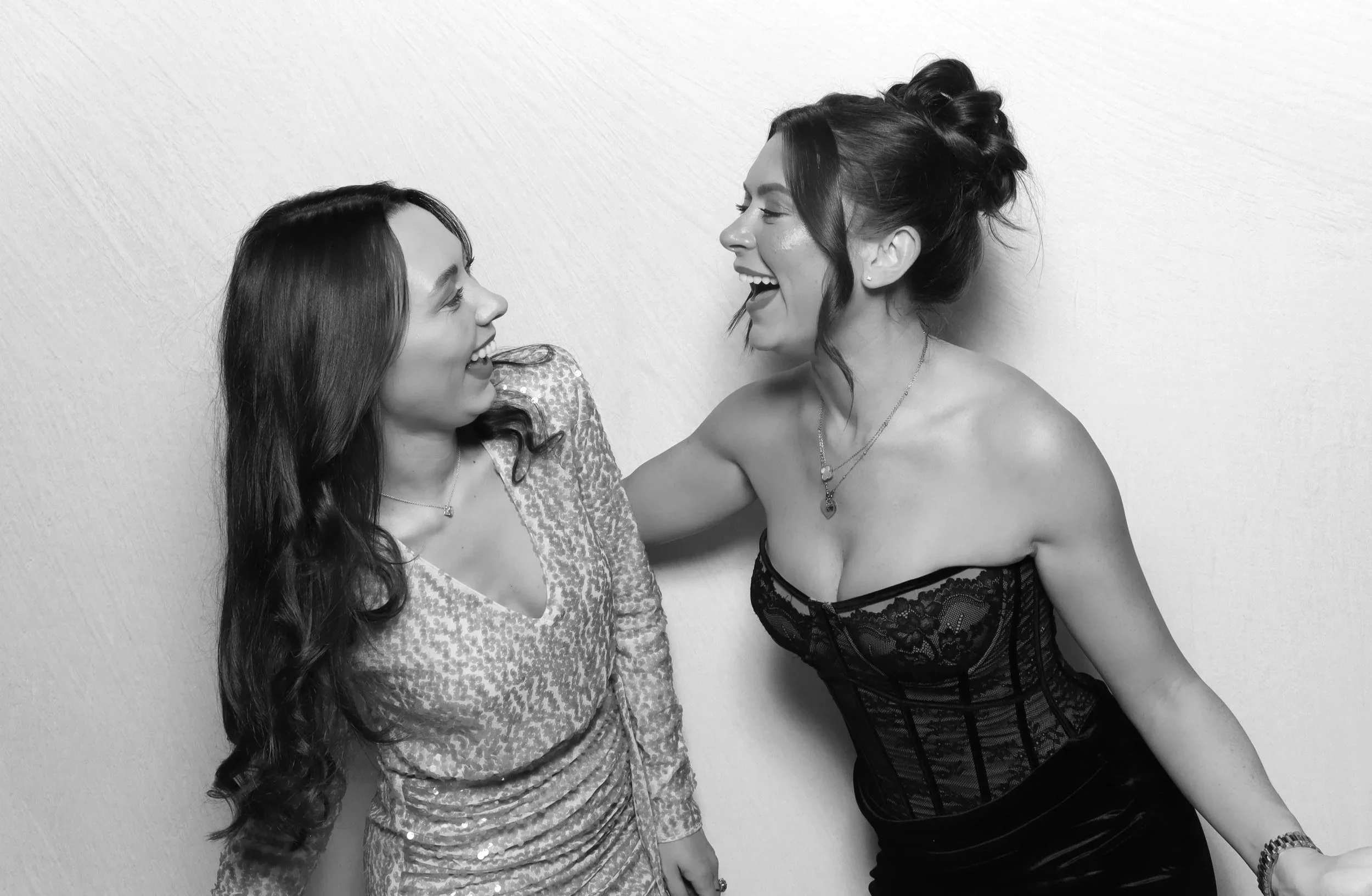 Two women with dark hair smiling and facing each other, one wearing a leopard print top and the other wearing a black lace top, against a plain white wall.