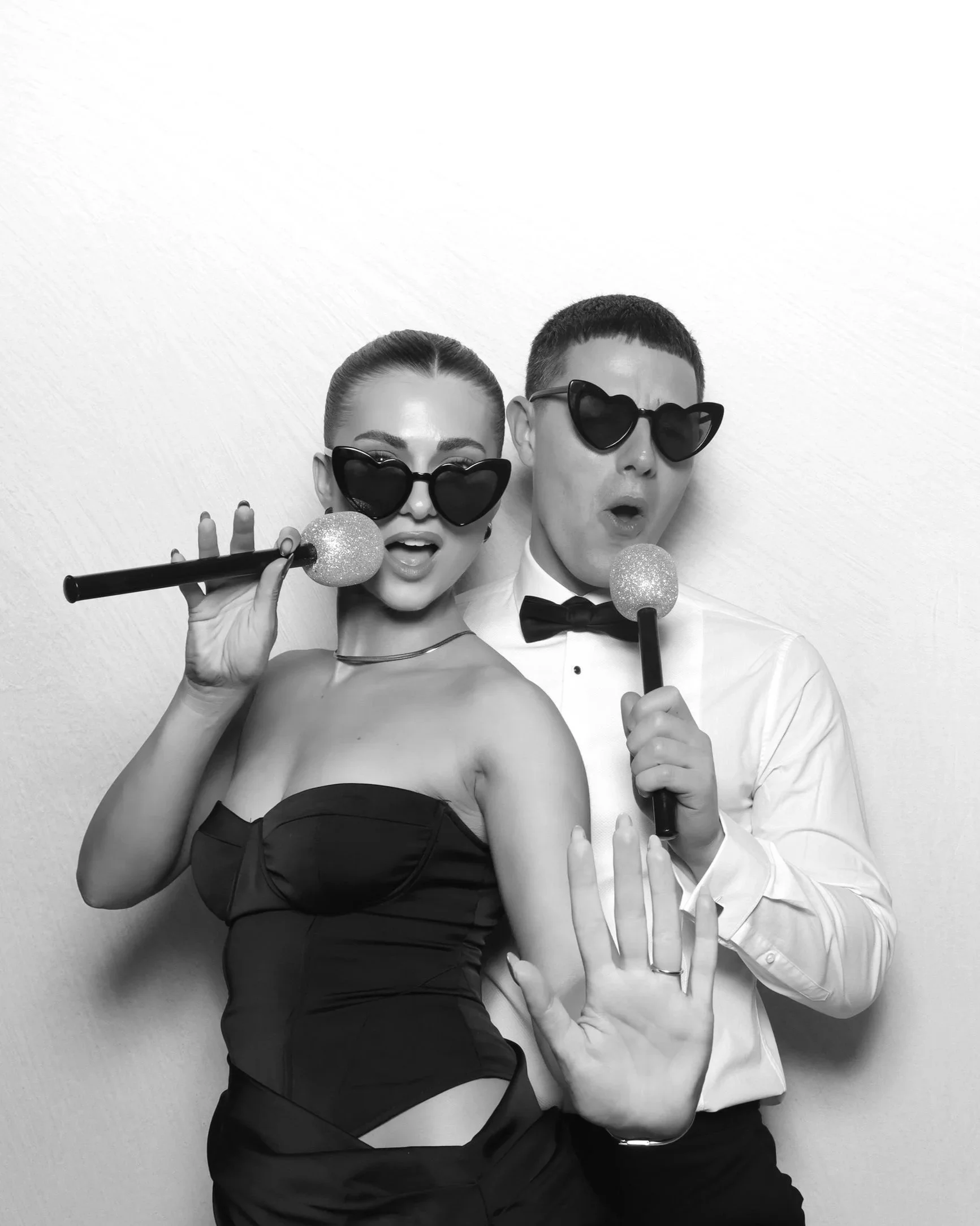 Black and white photo of a man and woman wearing sunglasses with heart-shaped lenses, posing with microphones and making playful gestures.