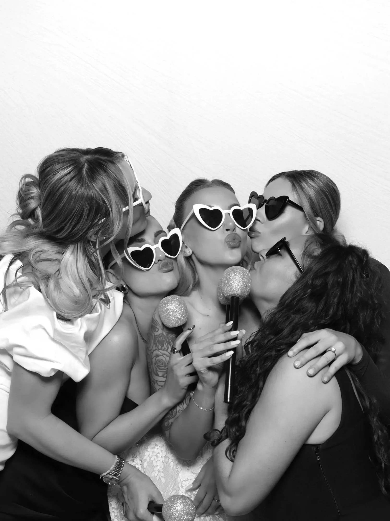 Group of women with party glasses and accessories, taking a photo together in a close, affectionate pose, holding two glittery microphones.