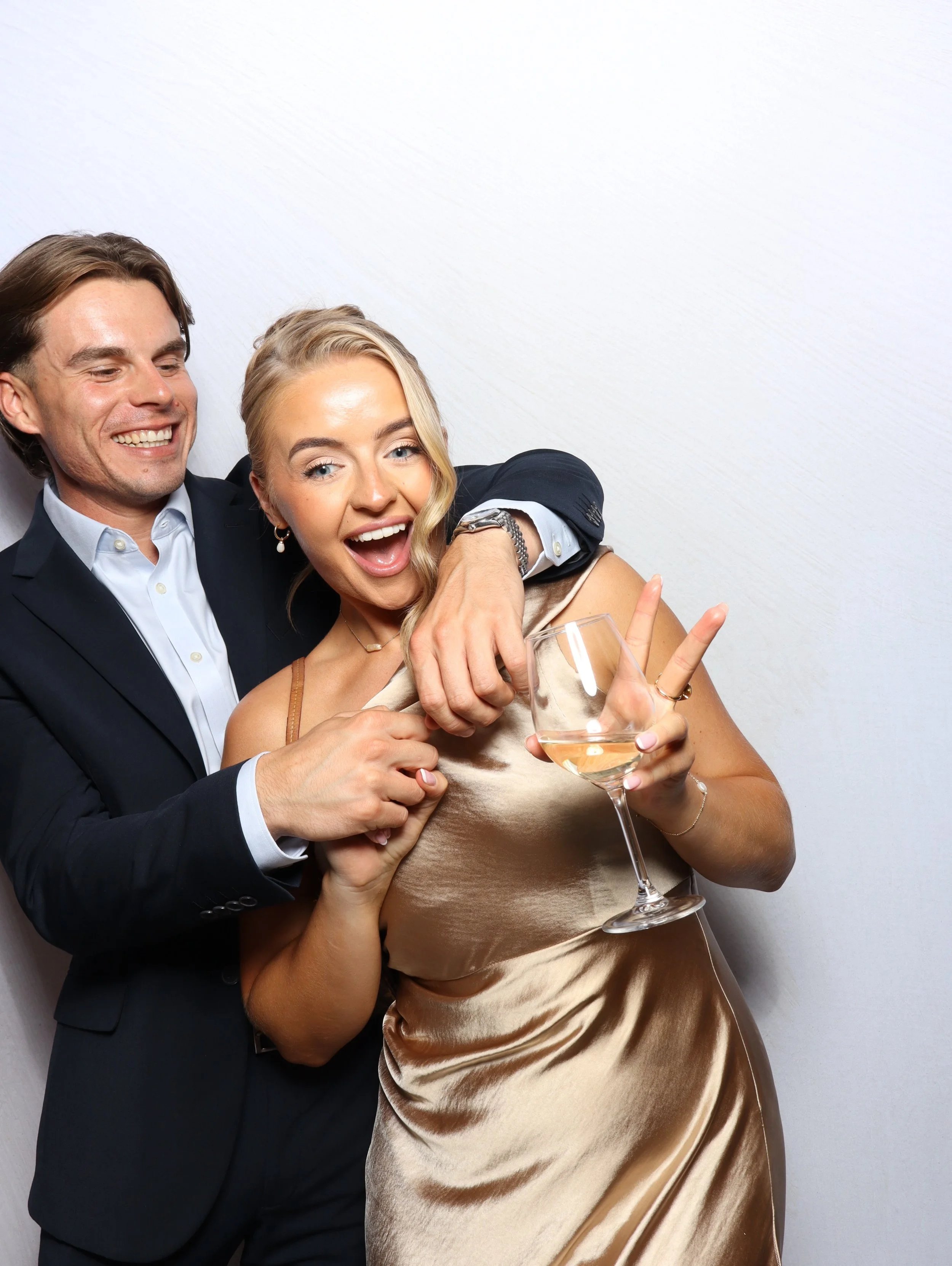 A man in a suit and a woman in a satin dress celebrate together, with the woman holding a glass of champagne and making a peace sign.