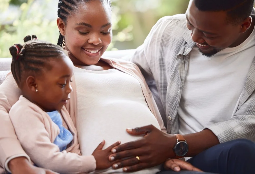 A family of three, including a pregnant woman, a young girl, and a man, sitting closely together and touching the woman’s pregnant belly.