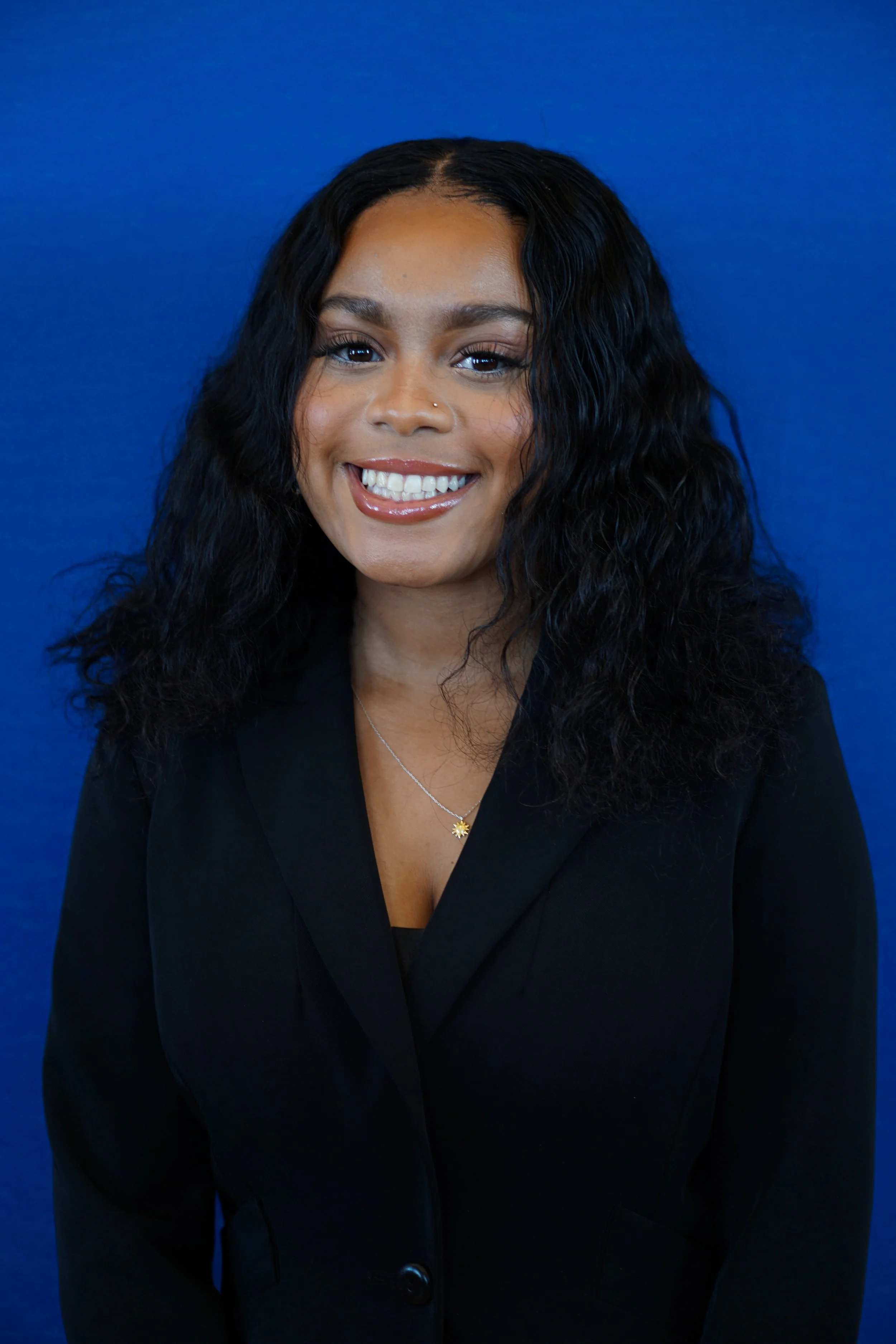A woman with dark, curly hair arranged in loose waves, smiling against a solid blue background. She wears a black blazer, a delicate necklace with a flower-shaped pendant, and has makeup with a focus on natural tones.