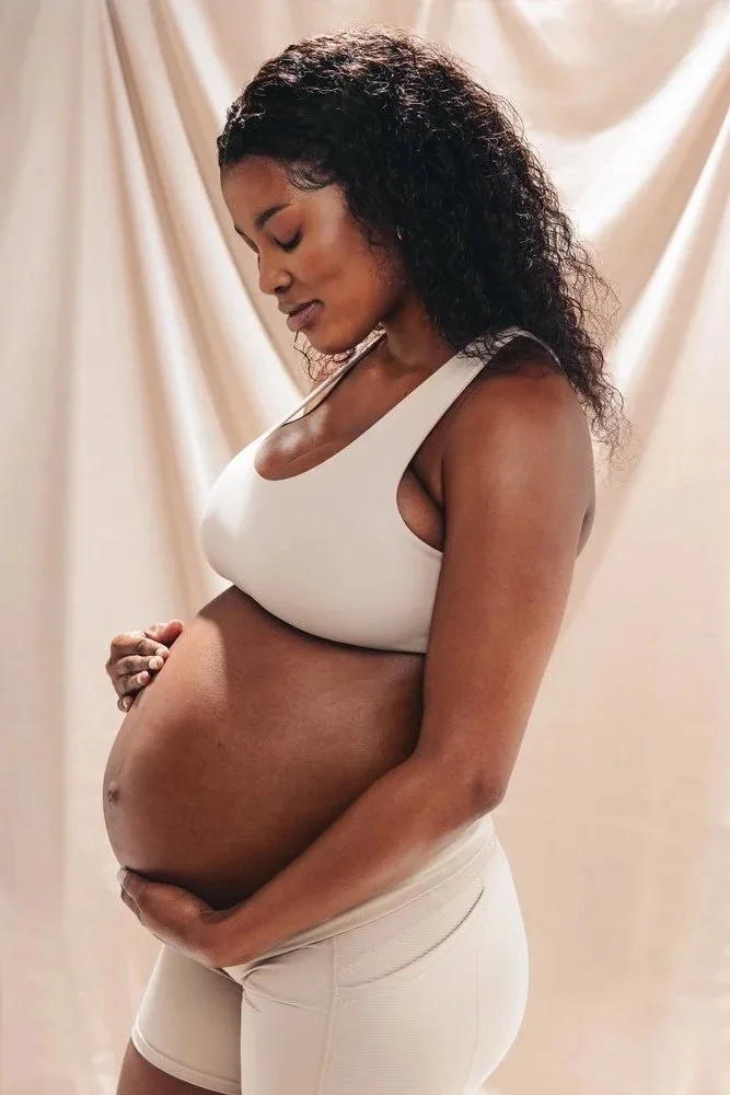 Pregnant woman with curly hair standing in front of a beige curtain, wearing a white sports bra and beige shorts, gently holding her belly with one hand, looking down with a serene expression.