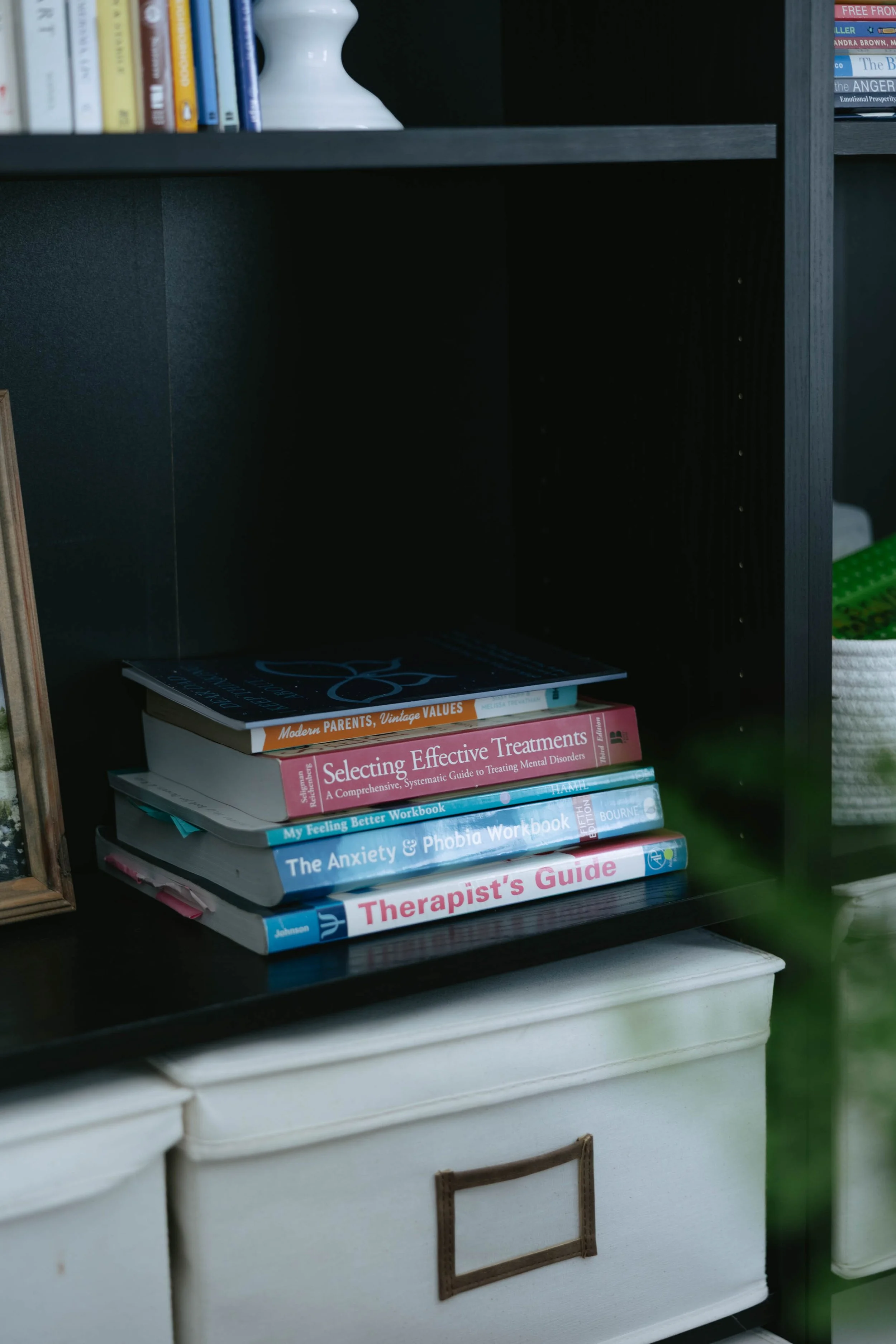 Books on topics related to mental health and therapy stacked on a dark bookshelf, with a white storage bin below.