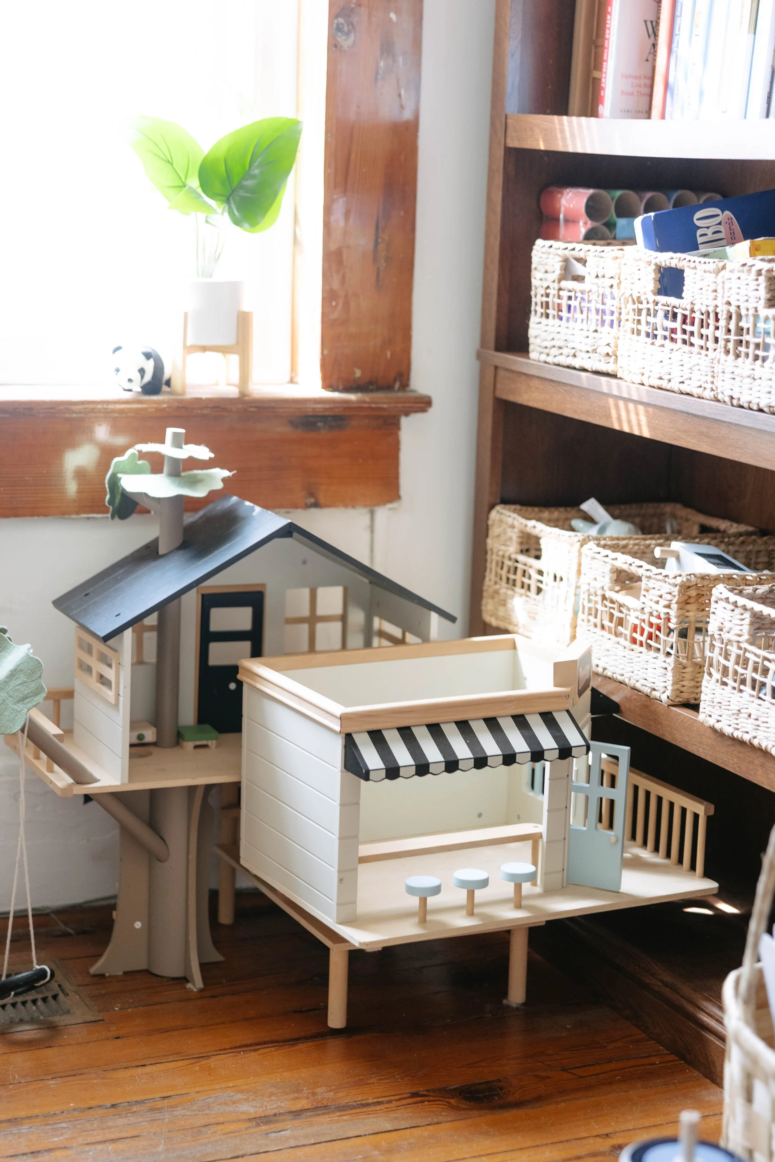 A children's dollhouse with outdoor patio furniture, a tree with four round green leaves, a small panda figurine, and a bookshelf with books and storage baskets in a cozy room with wooden floors.