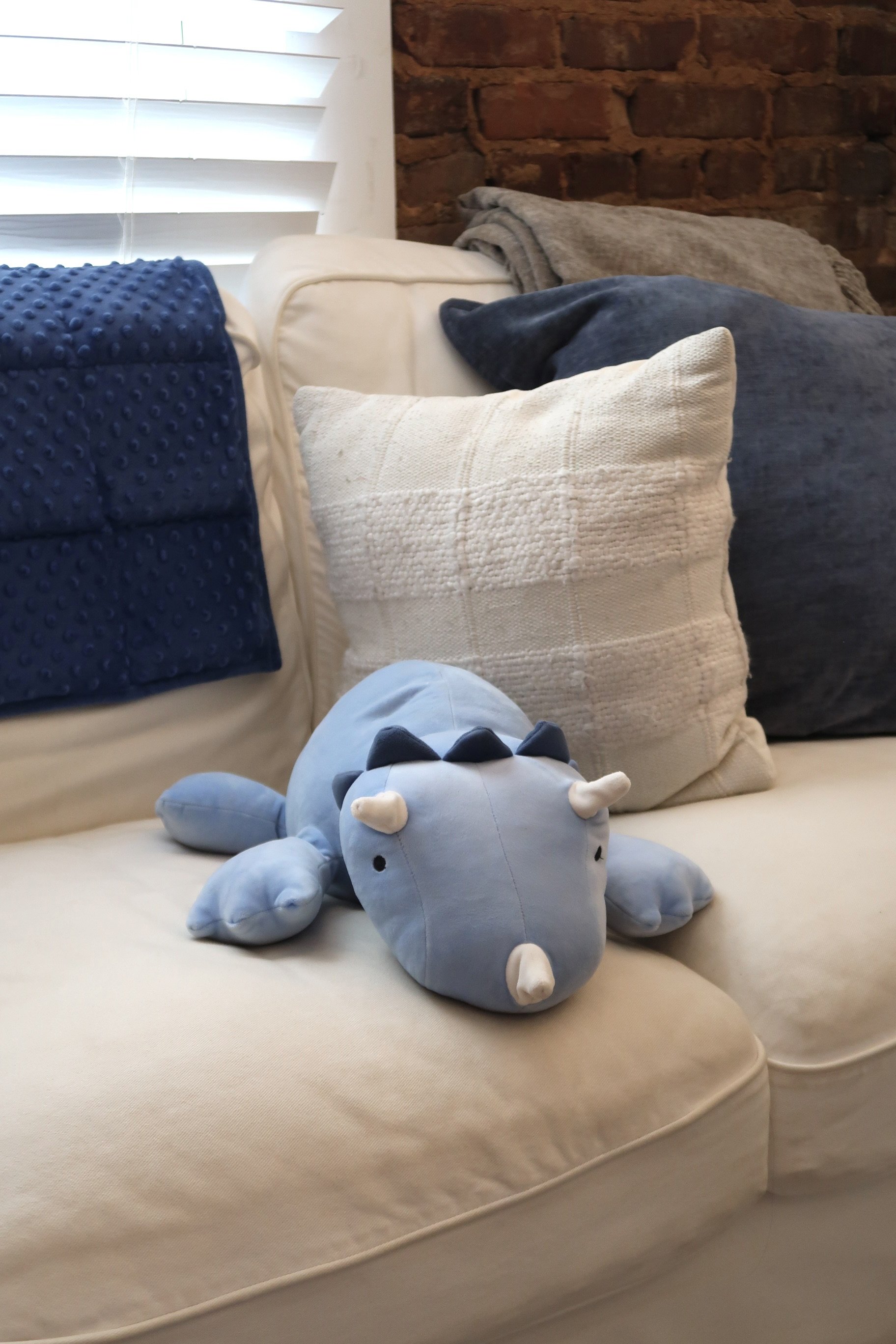 A plush blue rhinoceros toy with small white horns on a beige couch, with multicolored pillows and a brick wall in the background.