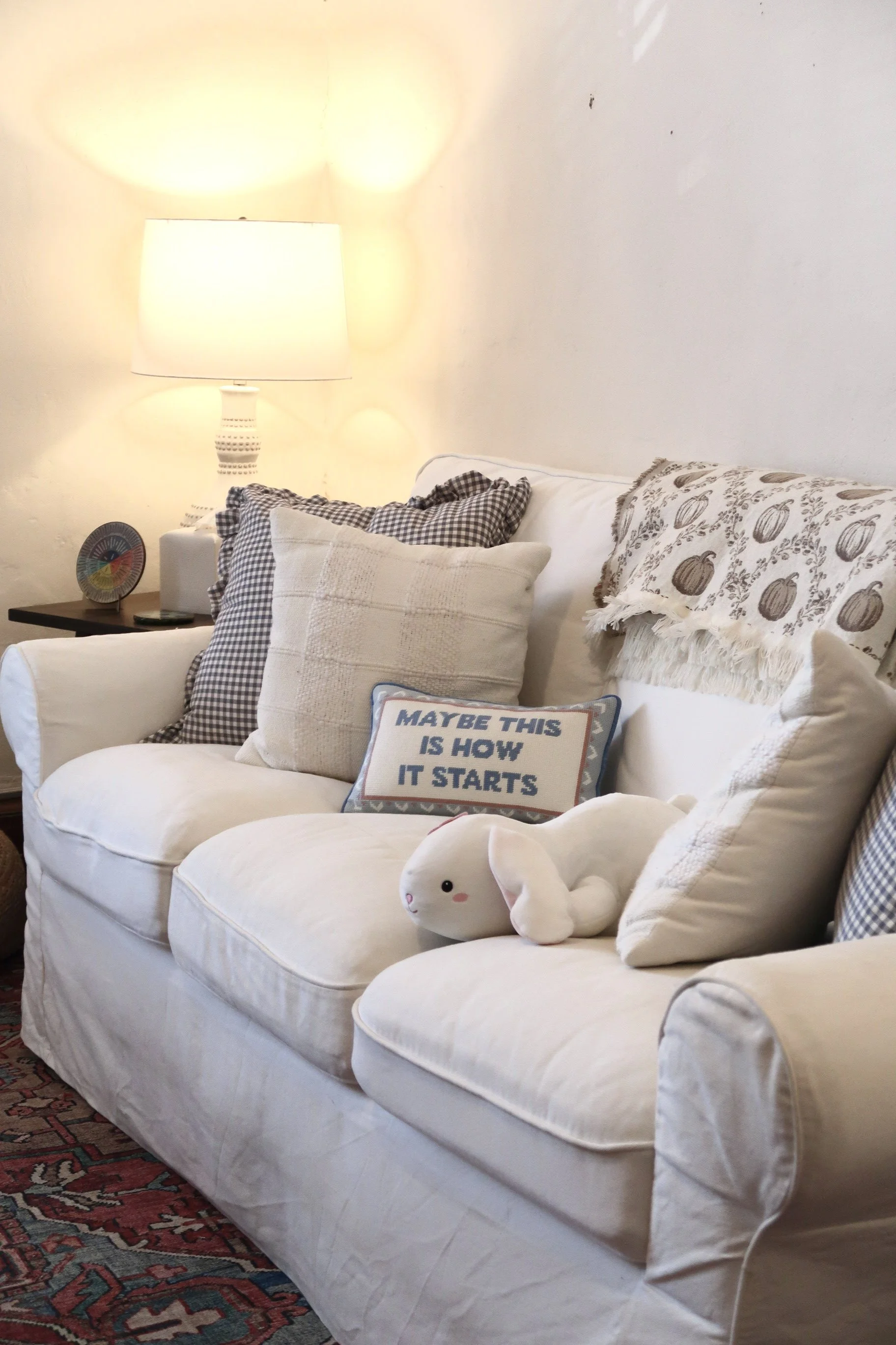 Office with a white sofa decorated with pillows, including one with a humorous message, a plush bunny toy, a side table with a closed fan and remote, and a lamp on the wall lighting the space.