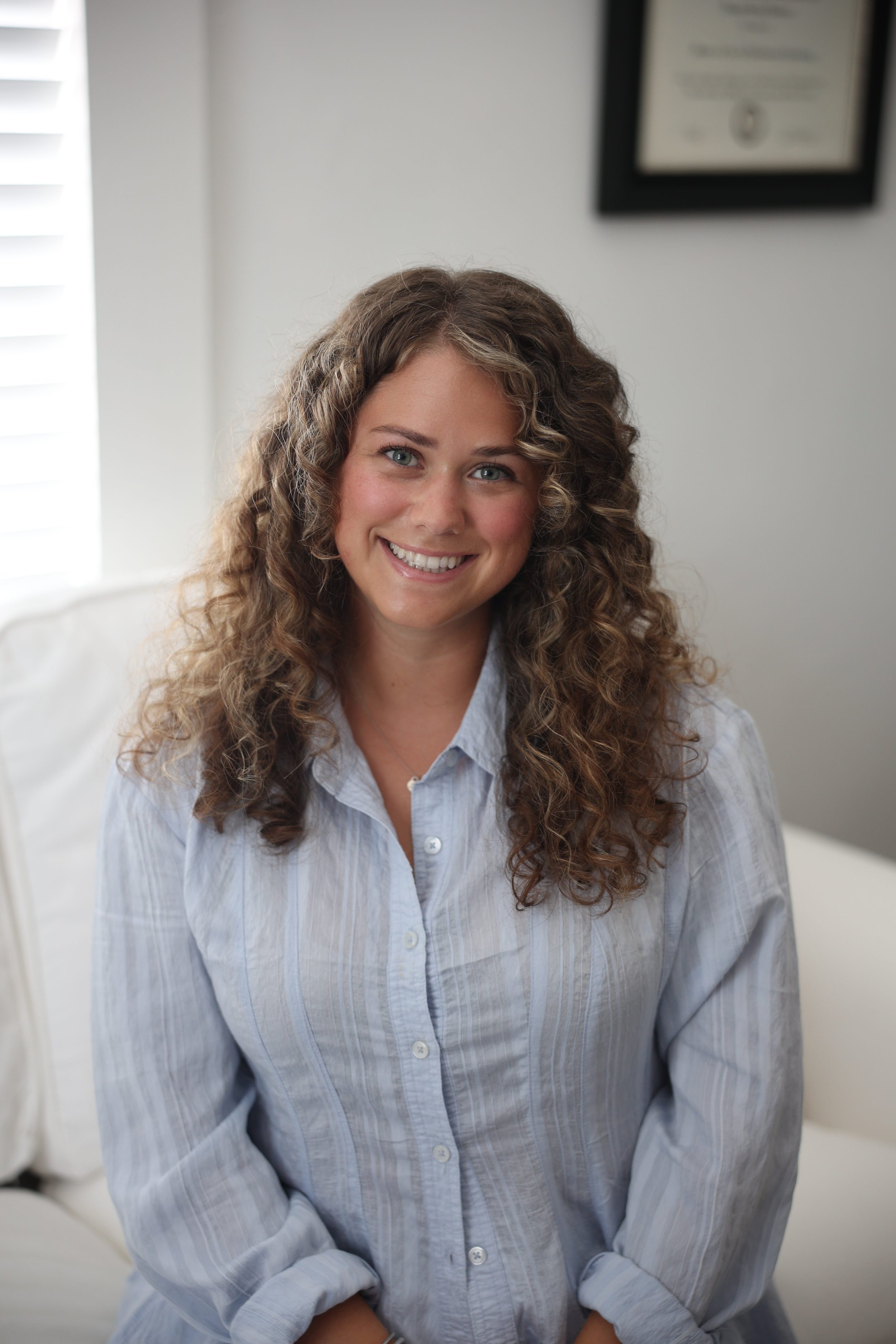 Smiling woman with curly brown hair, wearing a light blue striped button-up shirt, sitting on a sofa in a bright room.