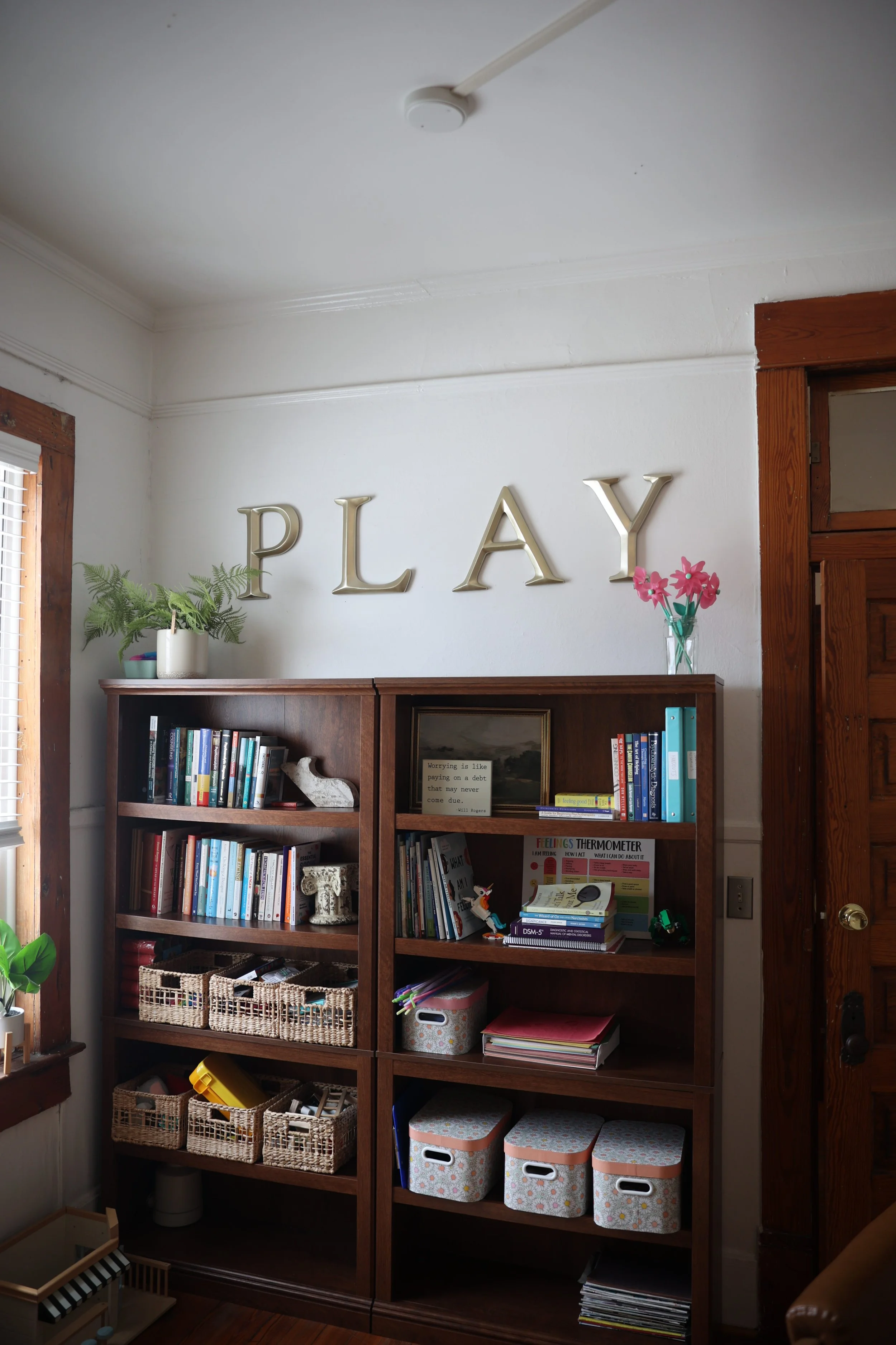 This image shows a wooden bookshelf filled with books, a framed quote, and decorative items. Above the bookshelf, the word 'PLAY' is displayed with large metallic letters on the wall. There are plants, baskets, and storage bins around the bookshelf, and a window with blinds illuminates the scene.