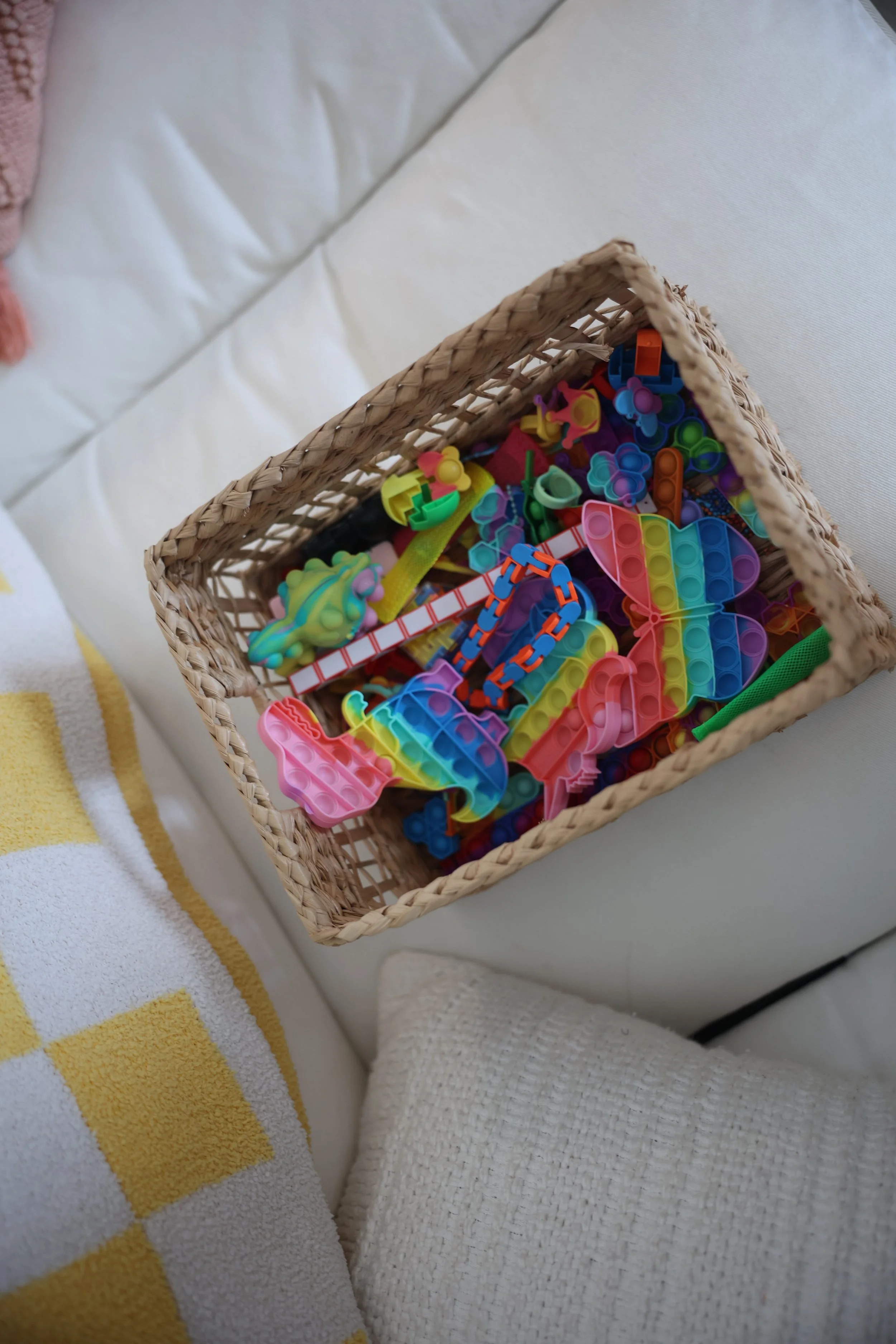 A woven basket filled with colorful plastic building toys is placed on a white surface near a bed with a white mattress and a yellow and white striped towel.