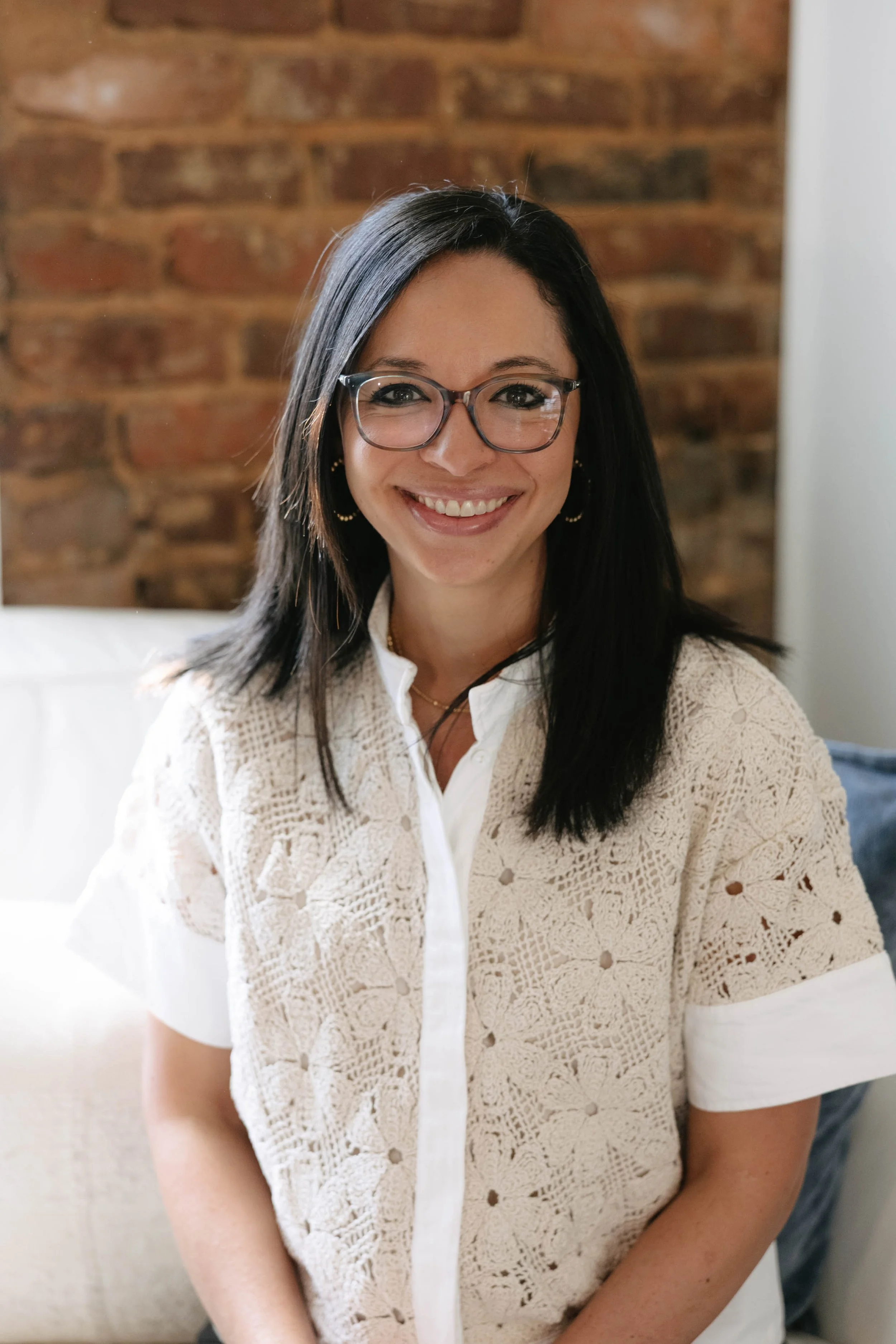A woman with long black hair, glasses, and hoop earrings smiling in front of a brick wall.