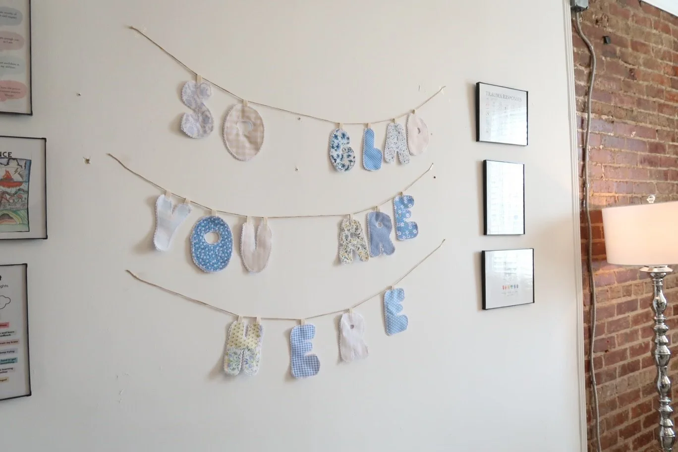 Decorative banner on a wall with the words 'so glad you are here' made from fabric-covered letters, and framed certificates or artwork on the adjacent wall. There is a lamp with a silver base and beige shade to the right.