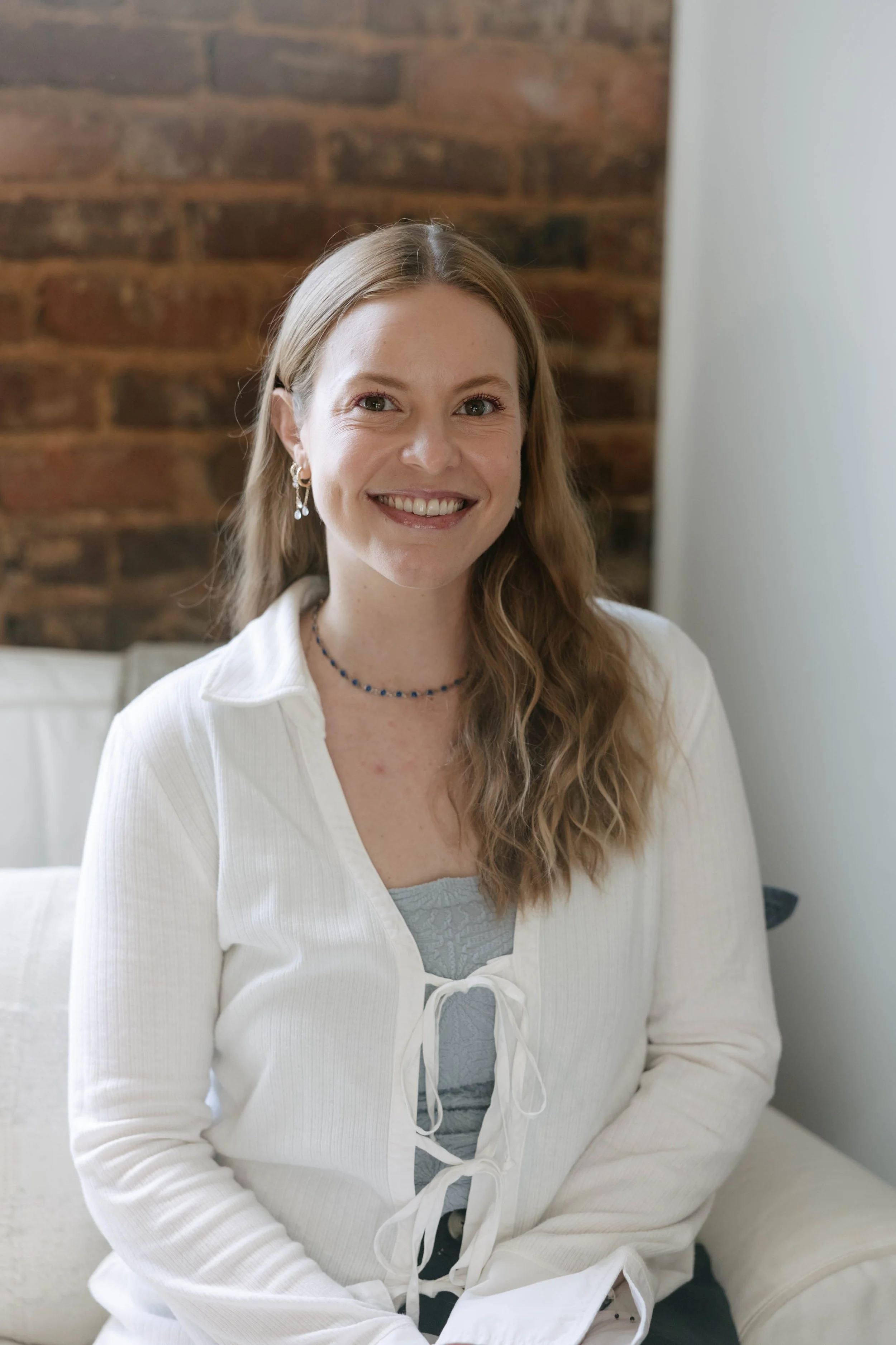 A woman with long wavy blonde hair, smiling, wearing earrings, a beaded necklace, a light blue top, and a white cardigan with ties, sitting indoors against a wooden brick wall and white furniture.