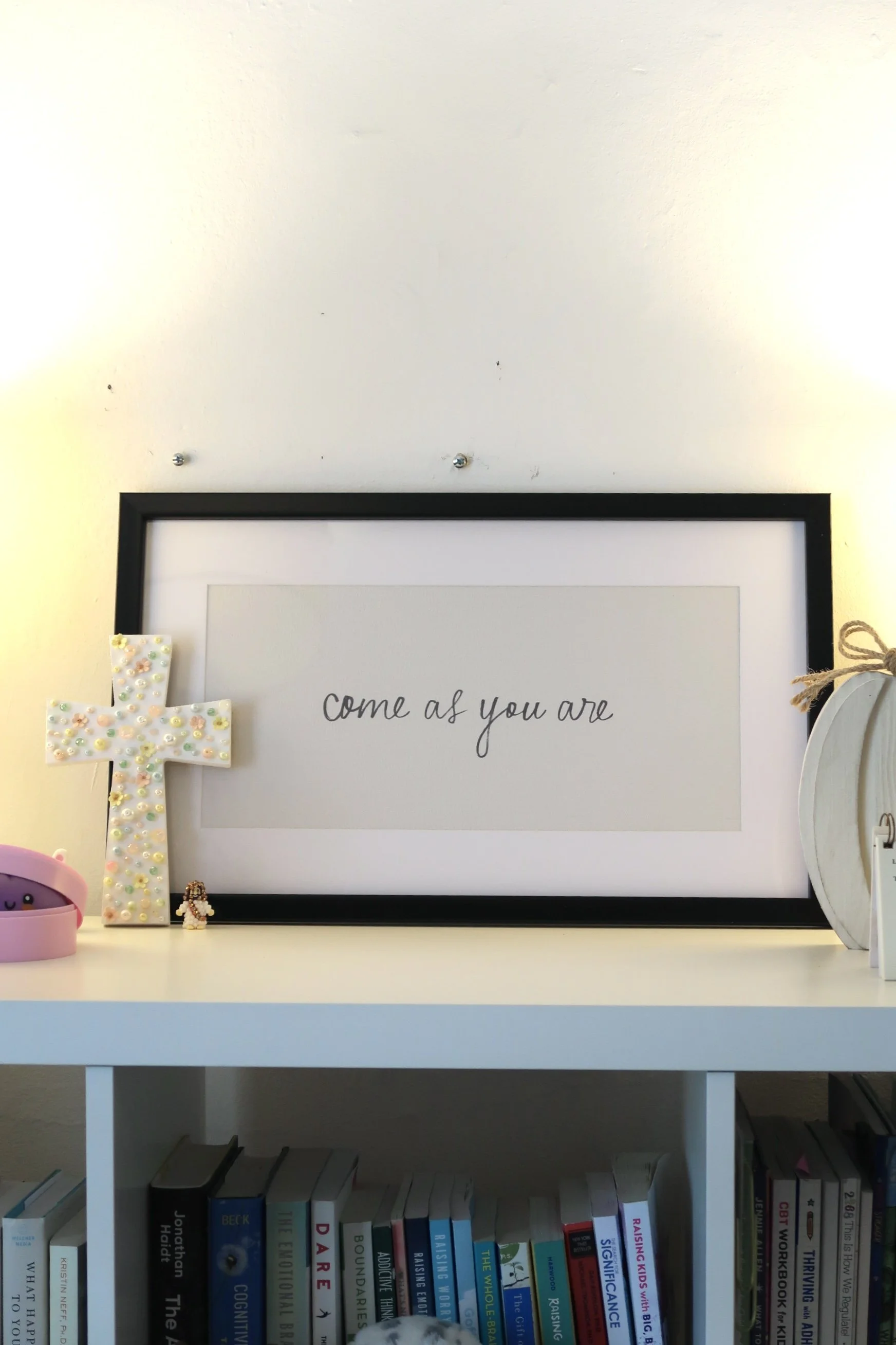 White-framed poster with the phrase 'come as you are' written in black cursive. Decorated with a cross made of pastel-colored beads on the left, a small figurine wearing a kimono, pink suction cup ears on a pink container, and books on a shelf below.