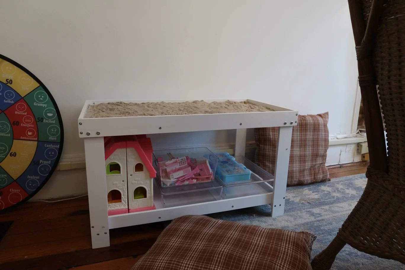 A white wooden table with a sandbox on top, filled with sand. Underneath, there are pink and clear plastic storage containers with small toys or objects. To the left, a part of a colorful wheel with various facial expressions is visible. There are brown plaid cushions on the floor and a wicker chair on the right side of the image.