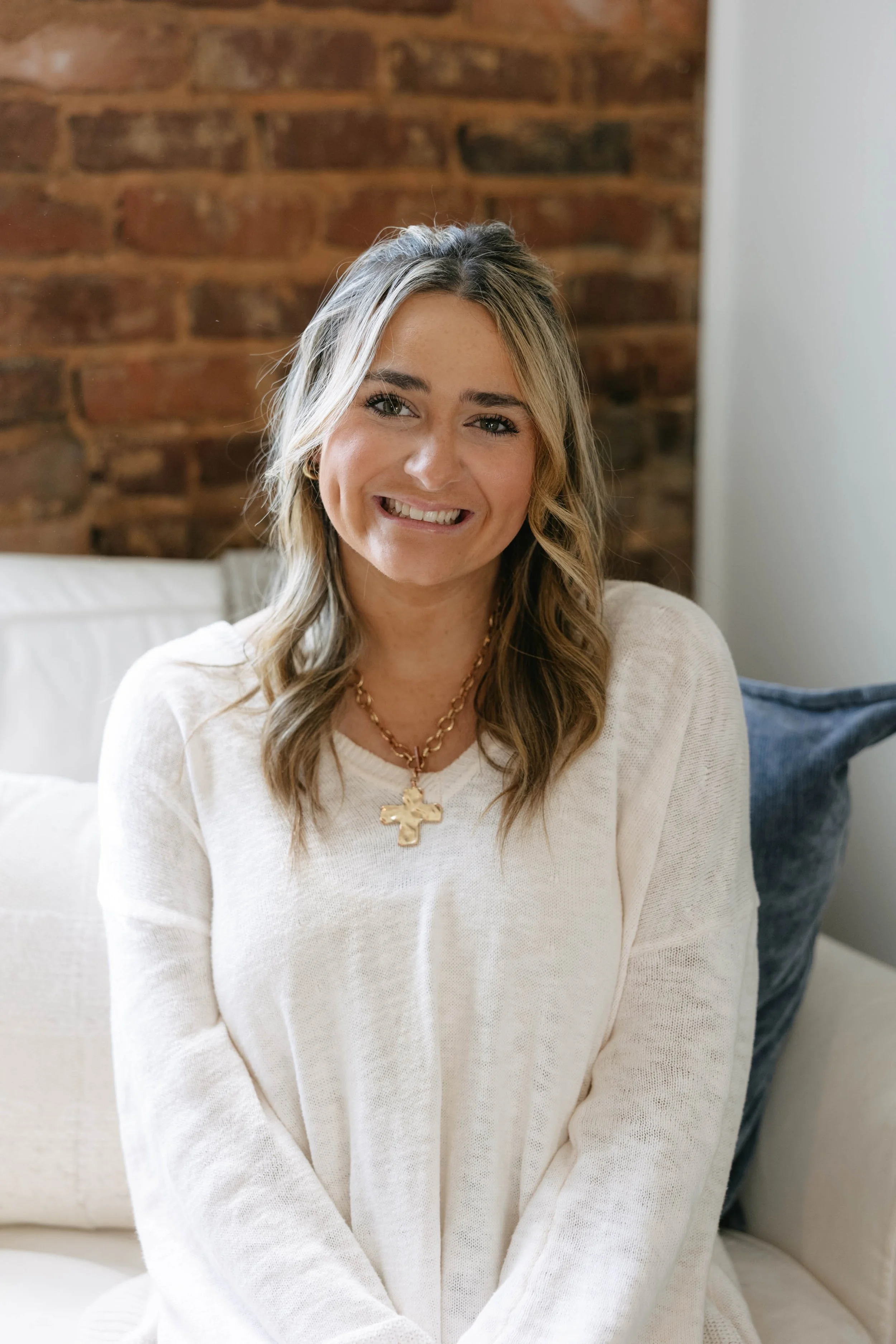 A woman with shoulder-length blonde hair smiling, wearing a white sweater and a gold cross necklace, sitting in a room with a brick wall background.