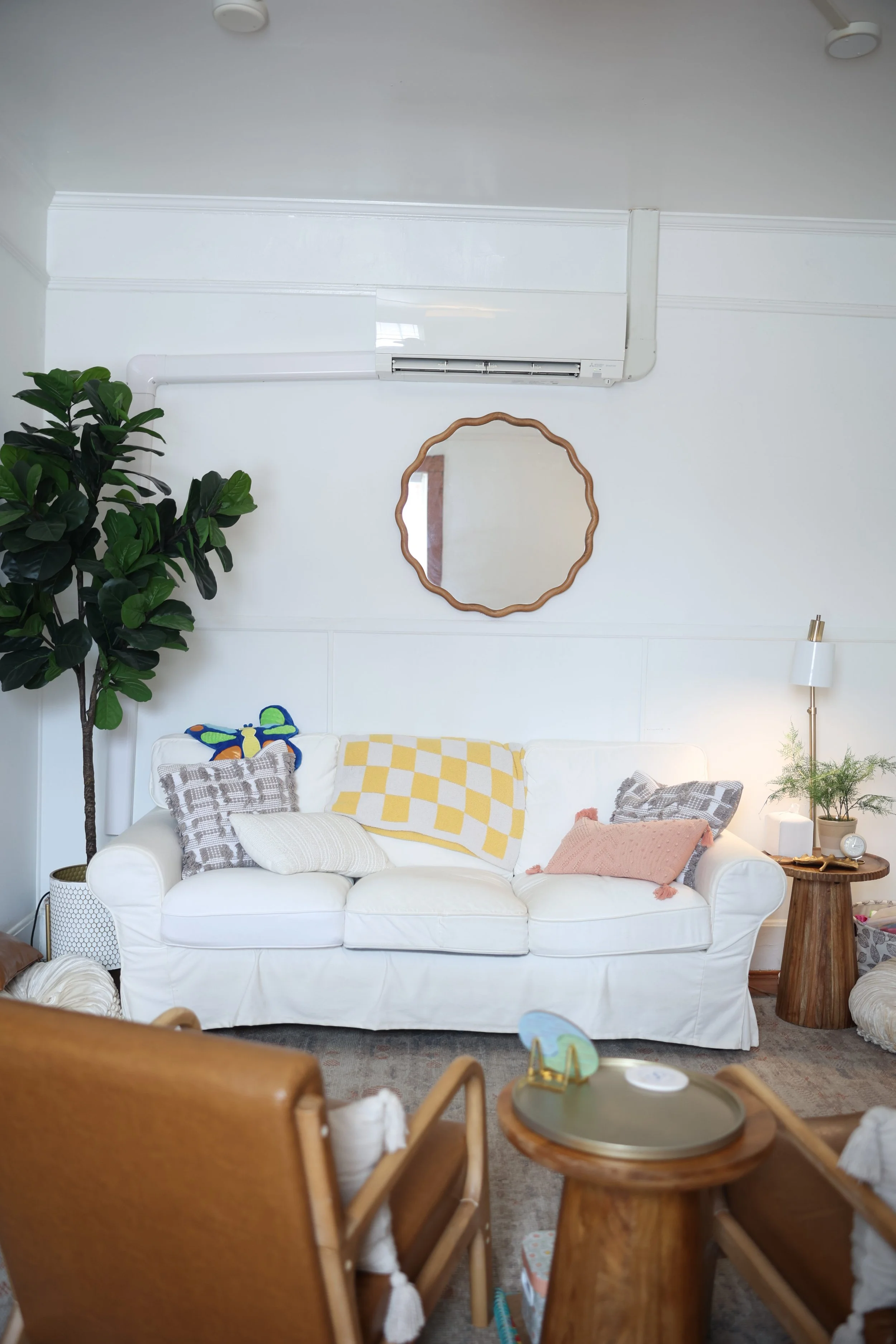 A cozy living room with a white couch decorated with various pillows and a yellow checkered blanket, a tall green indoor plant on the left, a round mirror on the white wall, a wooden side table with a lamp and clock, and a brown leather chair with a tray table in the foreground.