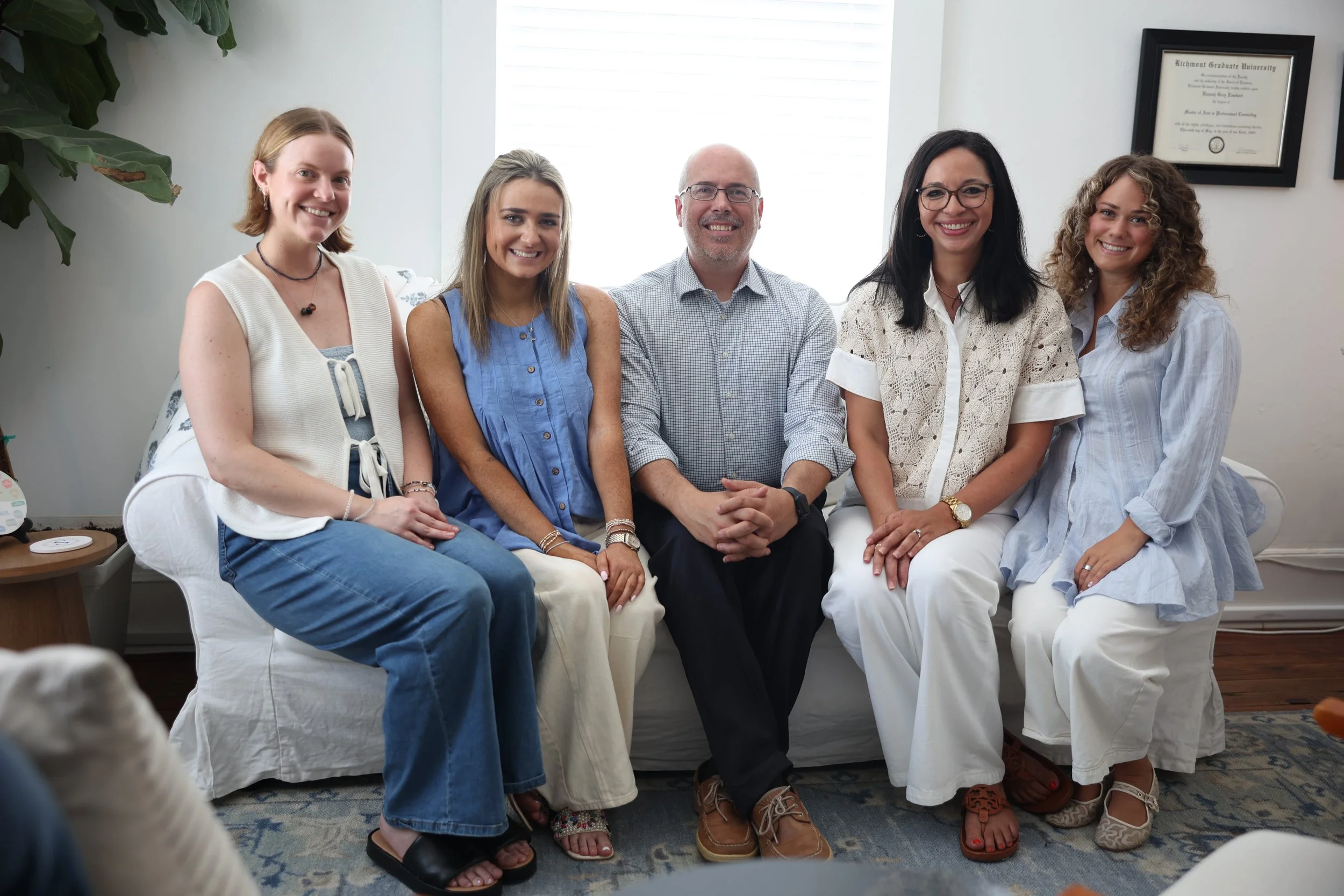 Group of five people sitting on a white couch, smiling at the camera, in a bright room with a framed diploma on the wall. There are large green plants and a window with white blinds in the background.