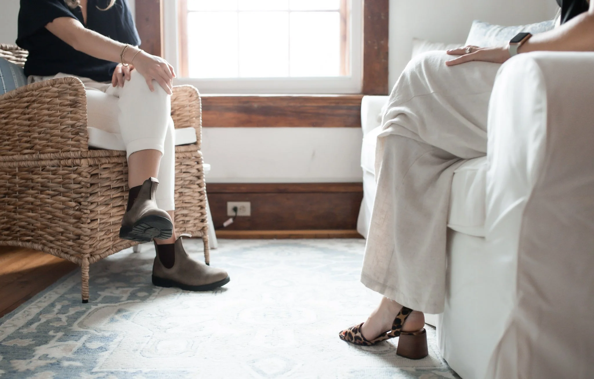 A woman sitting on a bed with her legs crossed, wearing leopard print high heels, and another person standing nearby wearing brown ankle boots, in a cozy room with natural light.