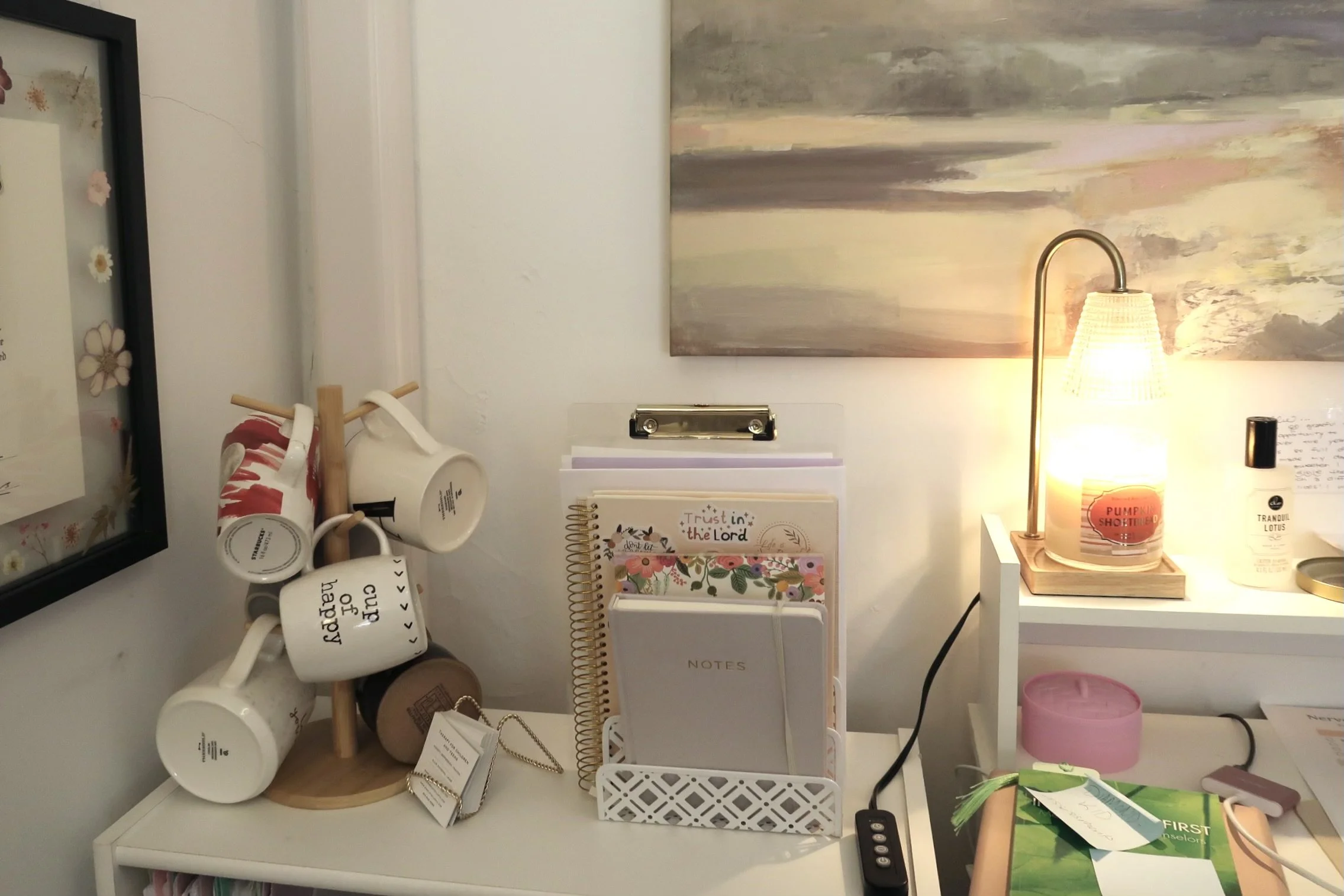 A desk with mugs, a wooden mug holder, a small lamp, notebooks, and various office supplies. There is a colorful abstract painting on the wall behind the desk.