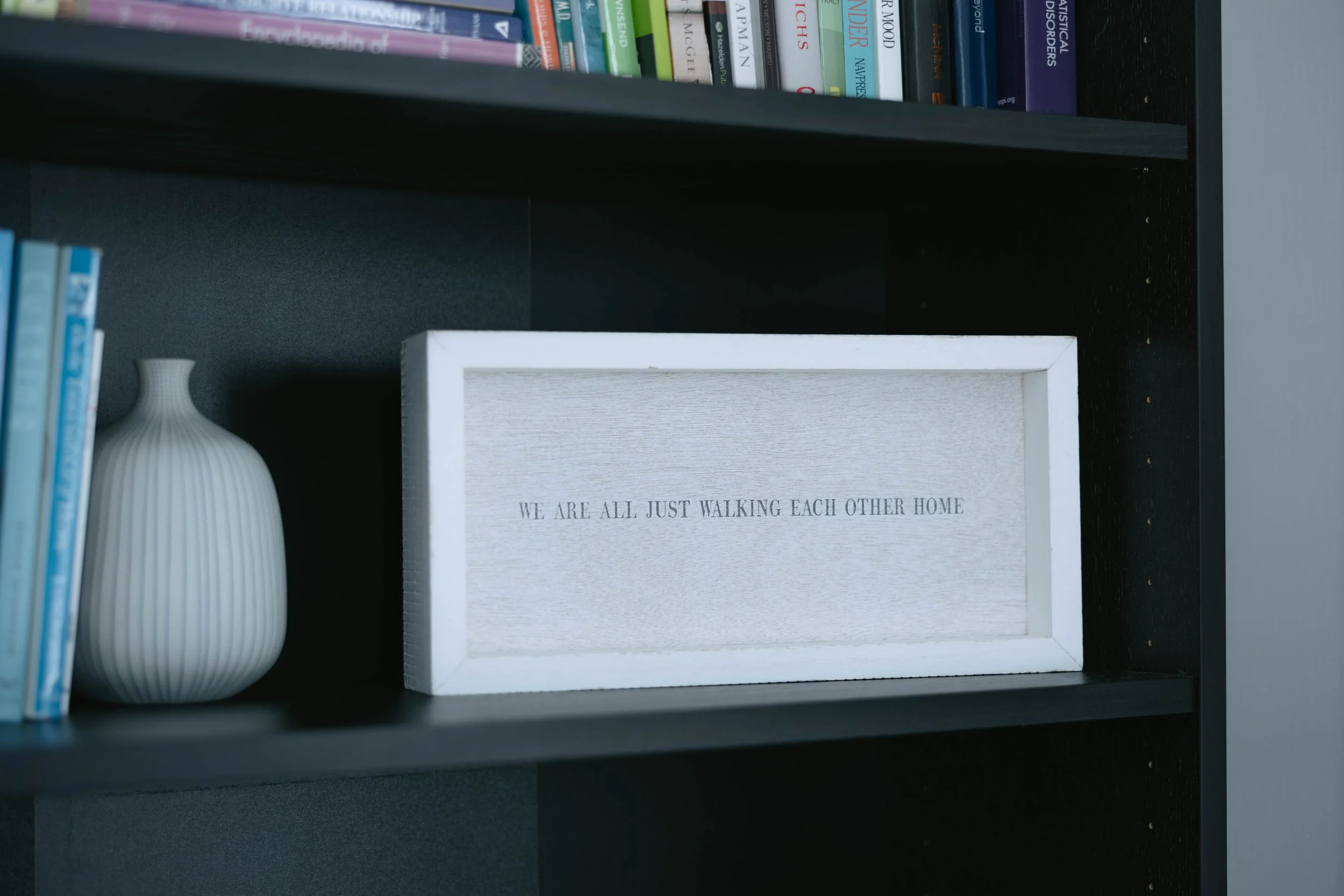 Close-up of a bookshelf with books, a white decorative vase, and a framed quote that says "We are all just walking each other home."