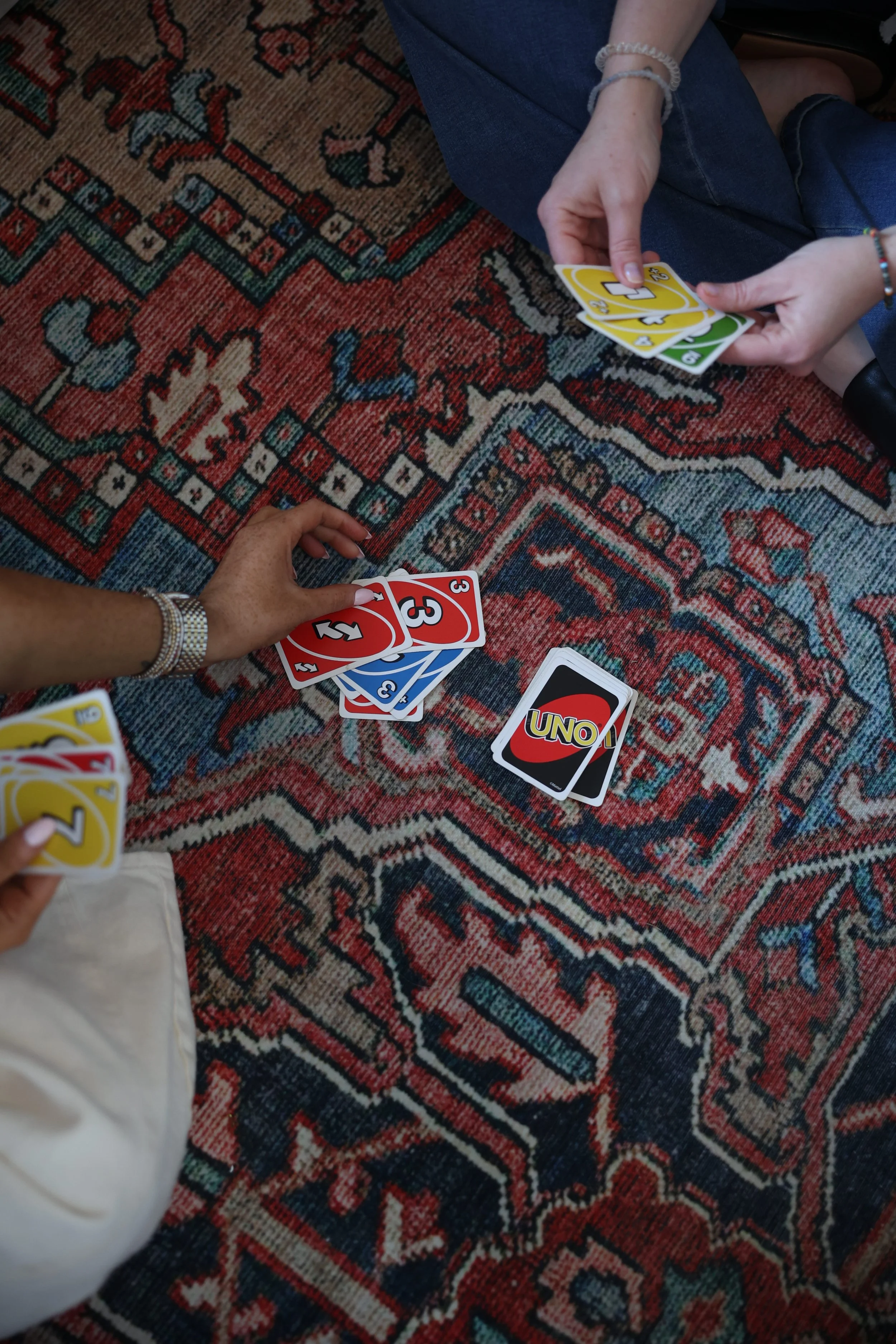 Two people are playing a card game on a patterned rug, with one person holding red and blue UNO cards and the other holding a yellow number card.