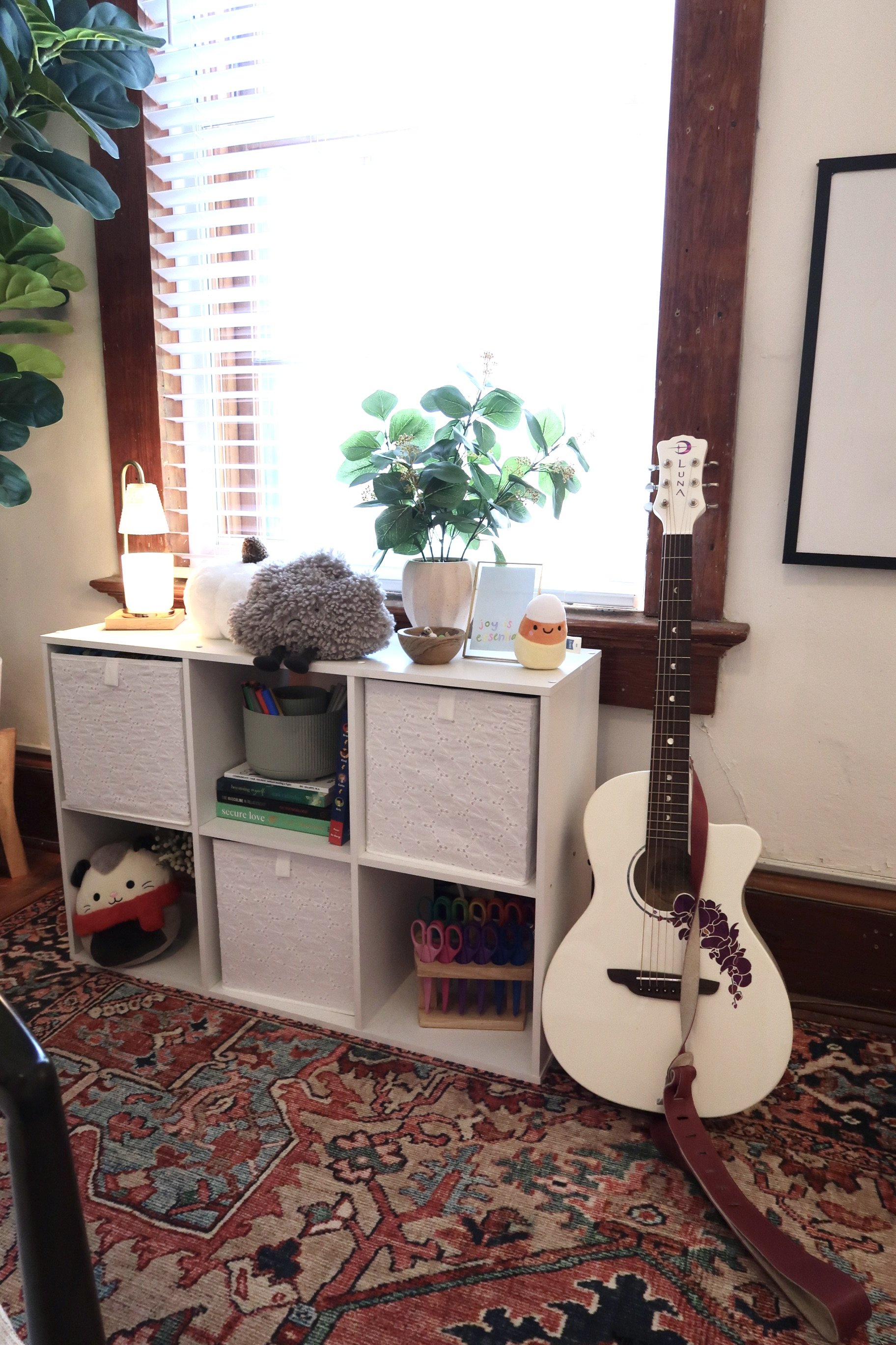 A white bookshelf with four compartments decorated with plush toys, books, and craft supplies, placed in front of a window with blinds, a wooden frame, and a dark red patterned rug on the floor. An acoustic guitar leans against the bookshelf.