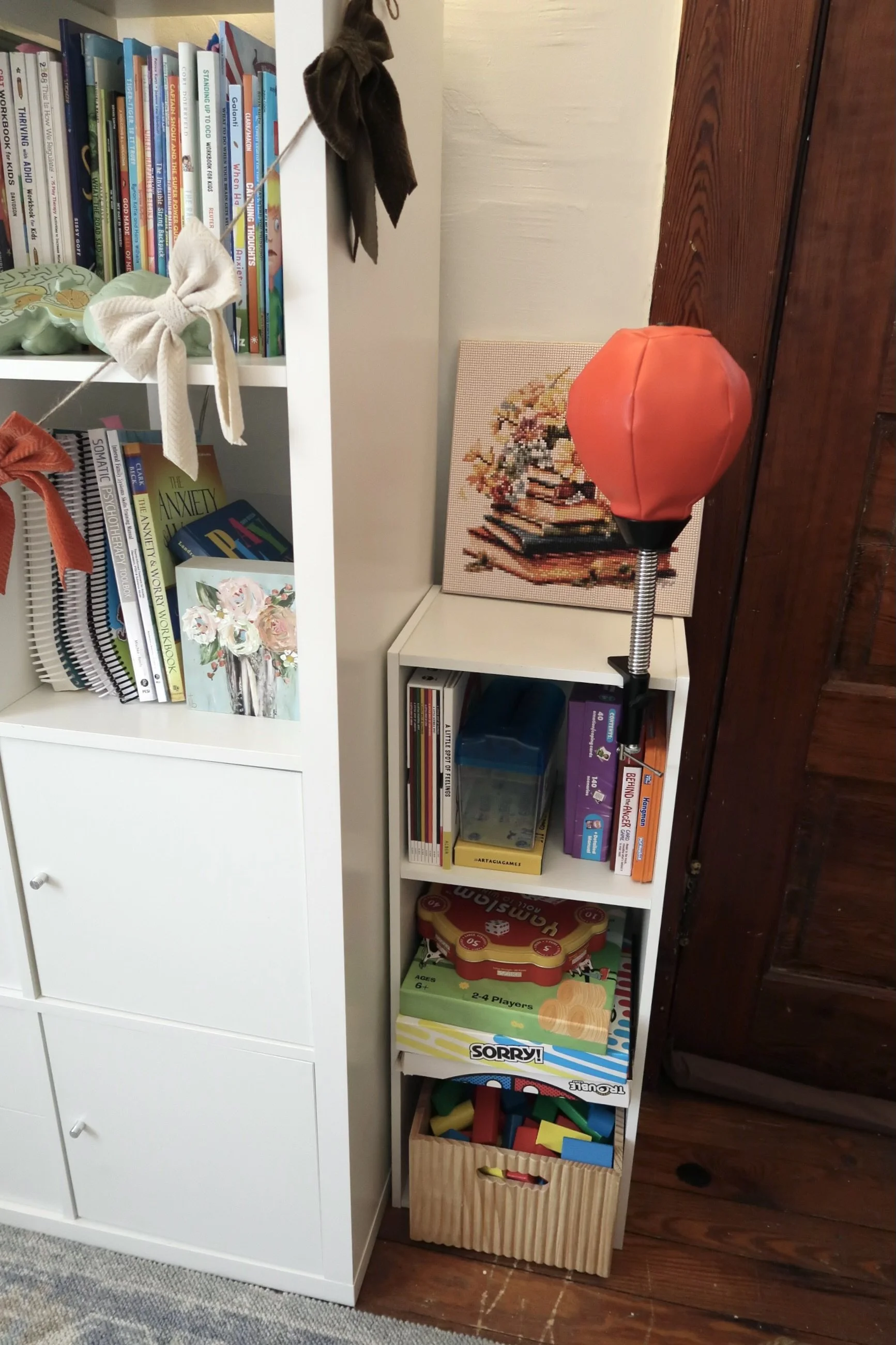 A white bookshelf with books, toys, and games, next to a small white shelf with table games, and a wooden basket filled with colorful building blocks, on a wooden floor.