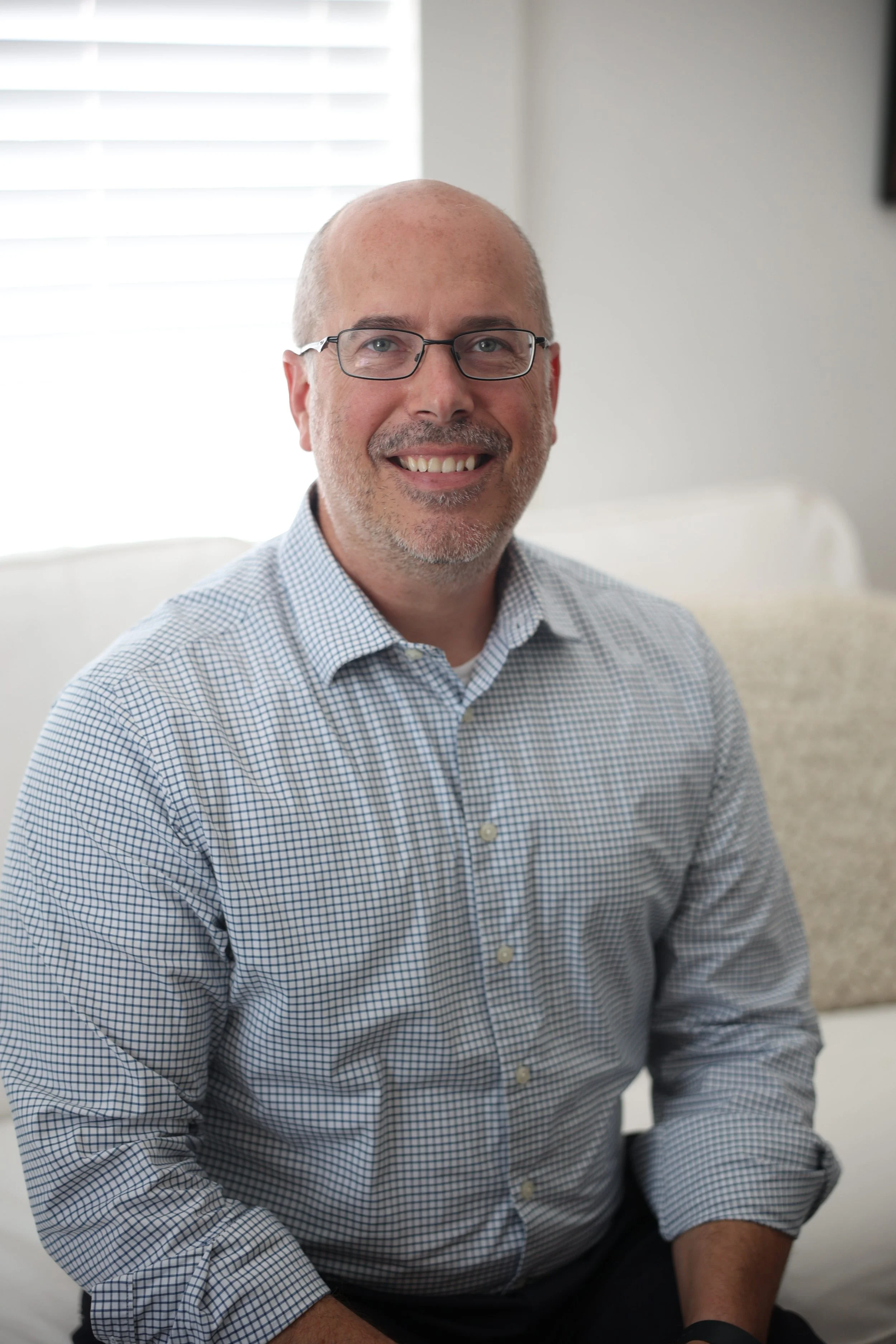 A middle-aged man with glasses, a short beard, and a bald head, smiling and wearing a light blue and white checkered shirt, sitting in a bright room with white blinds in the background.