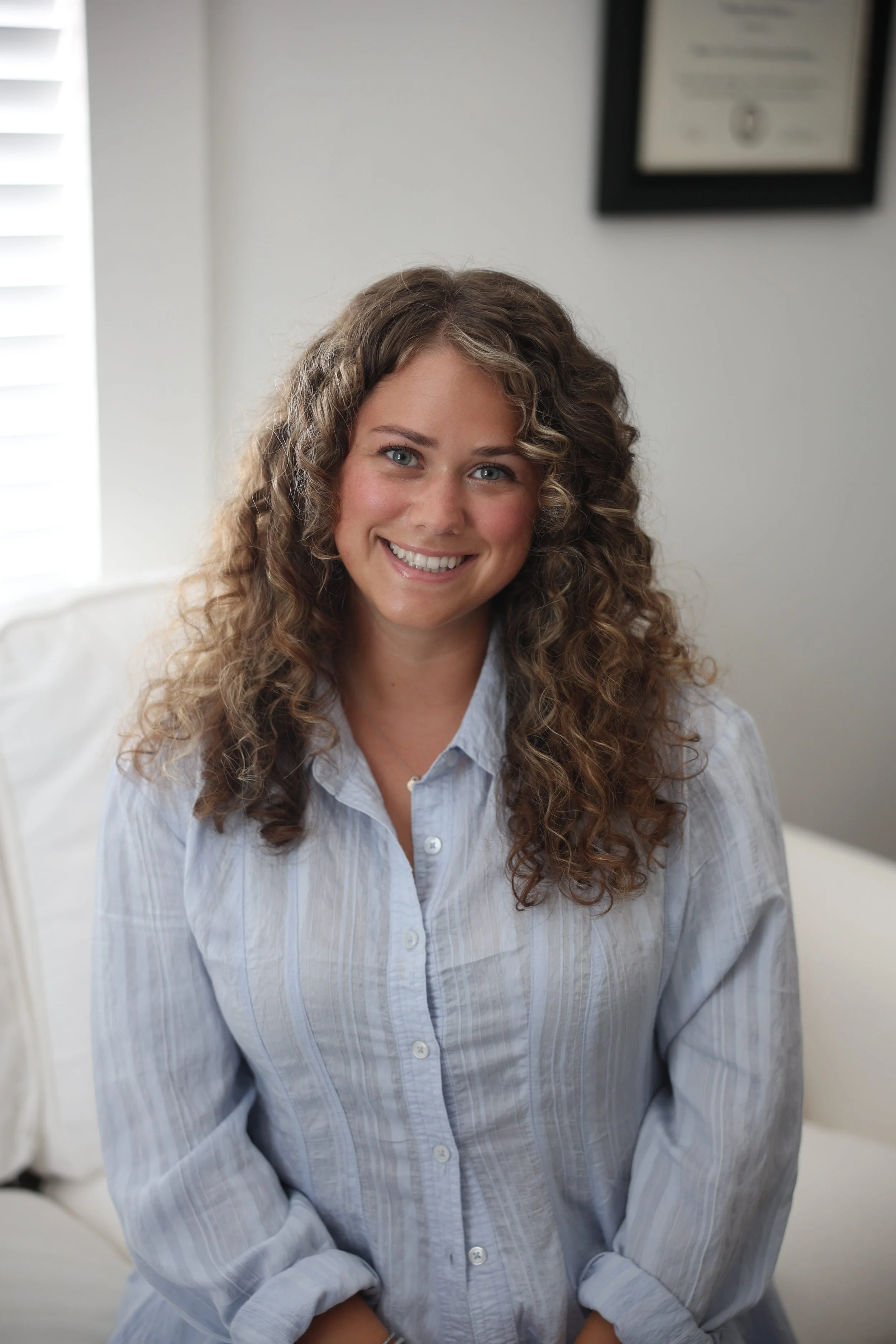 A young woman with curly brown hair and blue eyes, wearing a light blue button-up shirt, smiling at the camera. She is seated indoors with a blurred certificate or diploma hanging on the wall in the background.