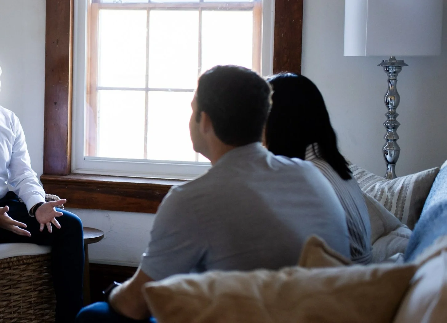 Three people sitting in a living room, engaged in conversation, with sunlight coming through a window behind them.