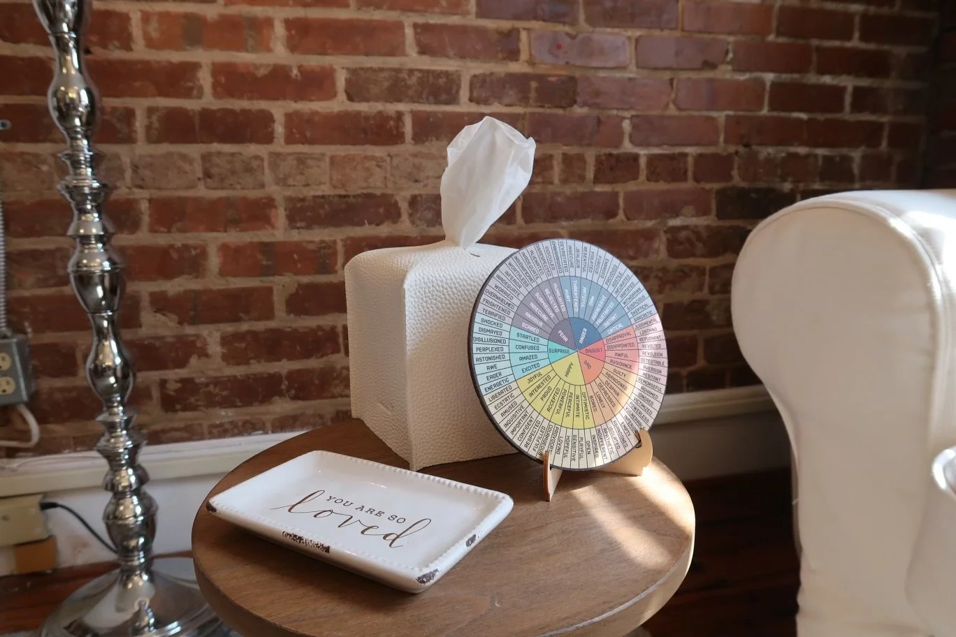 A tissue box with a decorative paper wheel on a wooden side table, a rectangular dish with the text 'You are so loved,' a shiny silver candlestick, and part of a white upholstered chair are visible against a brick wall background.