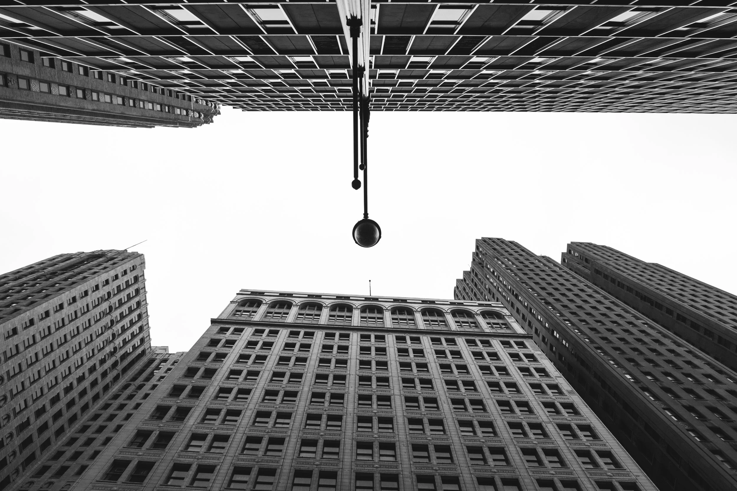 Black and white photo taken from below of several tall skyscraper buildings in an urban cityscape with intersecting and aligned windows, and a lamp post in the center.