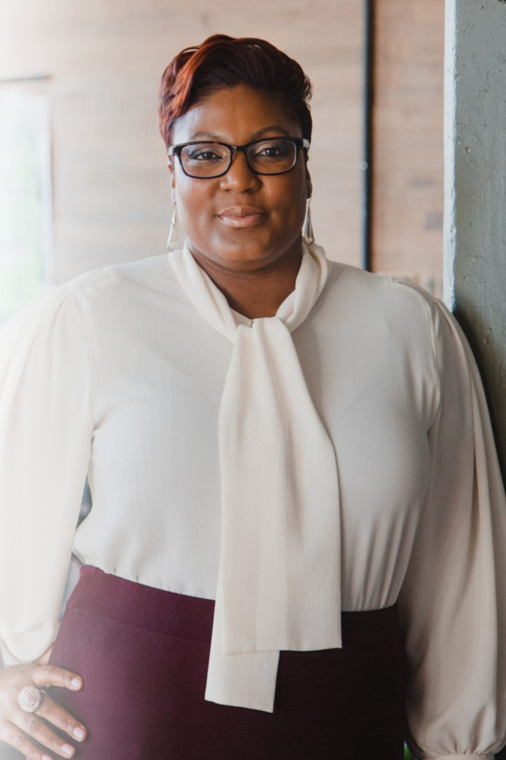 A woman with short reddish hair, glasses, and earrings, wearing a cream blouse with a bow and a maroon skirt, standing indoors near a wall.