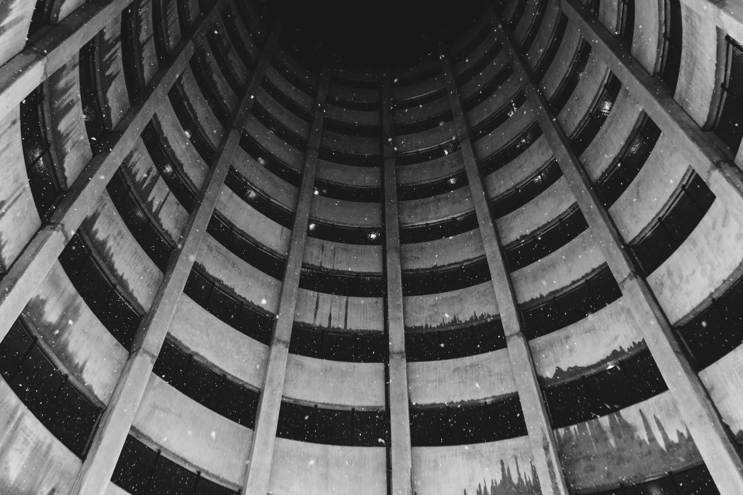 Looking up into the atrium of a tall, multi-story concrete building with repetitive, curved balconies on each floor. It appears to be taken at night, with some lights on inside, and snow falling in the scene.