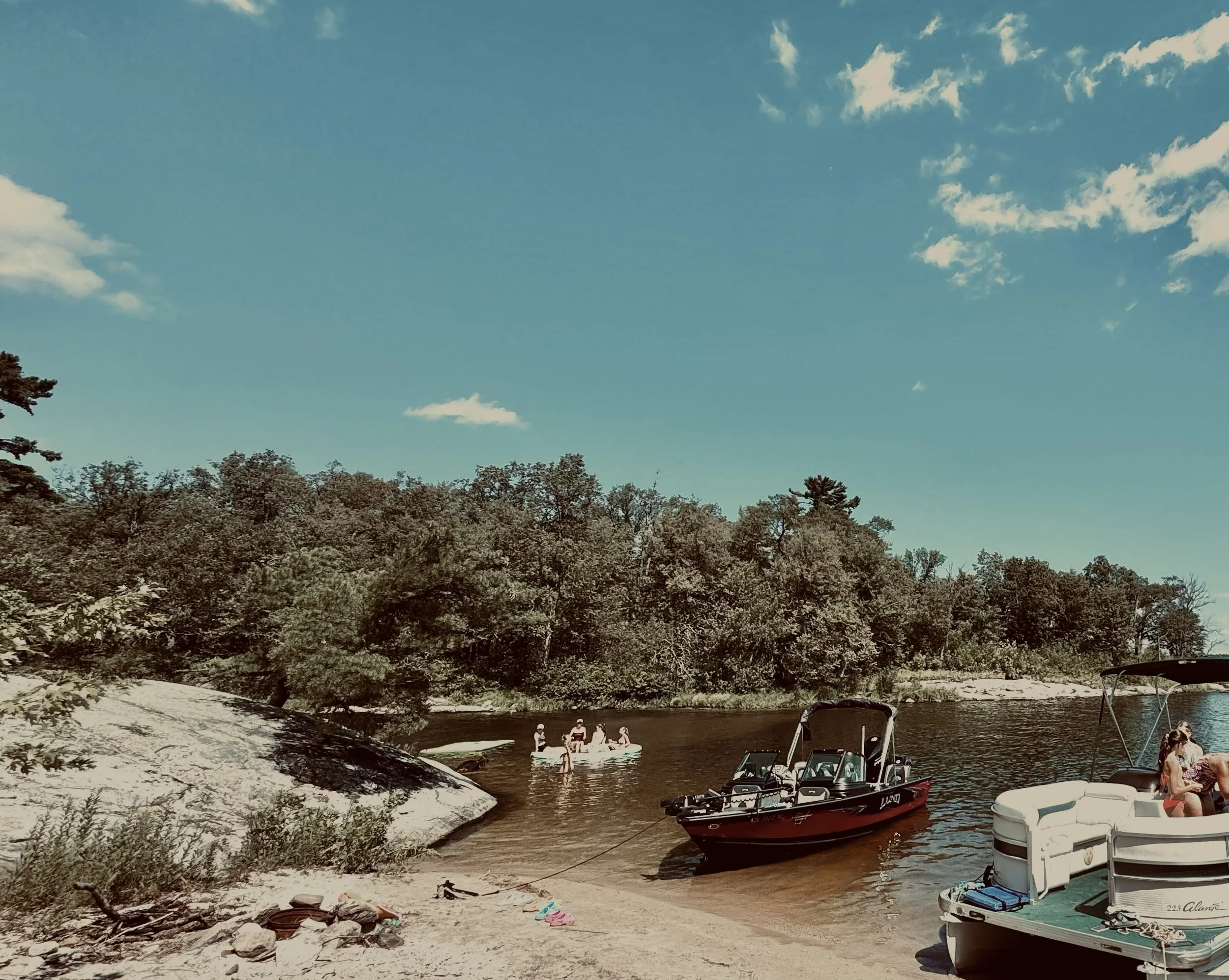 People enjoying a day on the water by a lakeside with boats, trees, and a rocky shoreline under a partly cloudy sky.