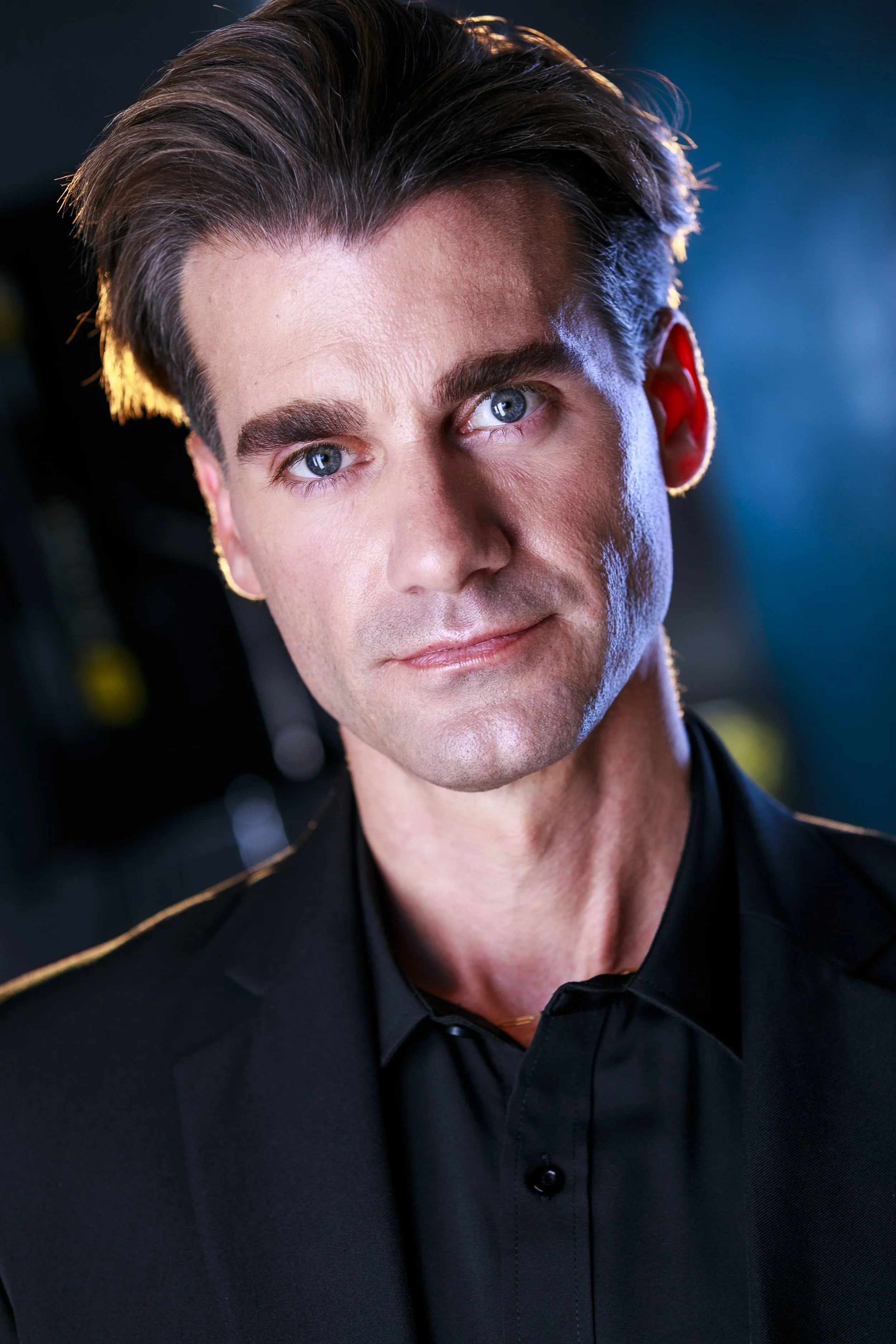 Close-up of a man with styled brown hair, blue eyes, and a light complexion wearing a black shirt with a gold accent on the collar, against a dark background with blue lighting.