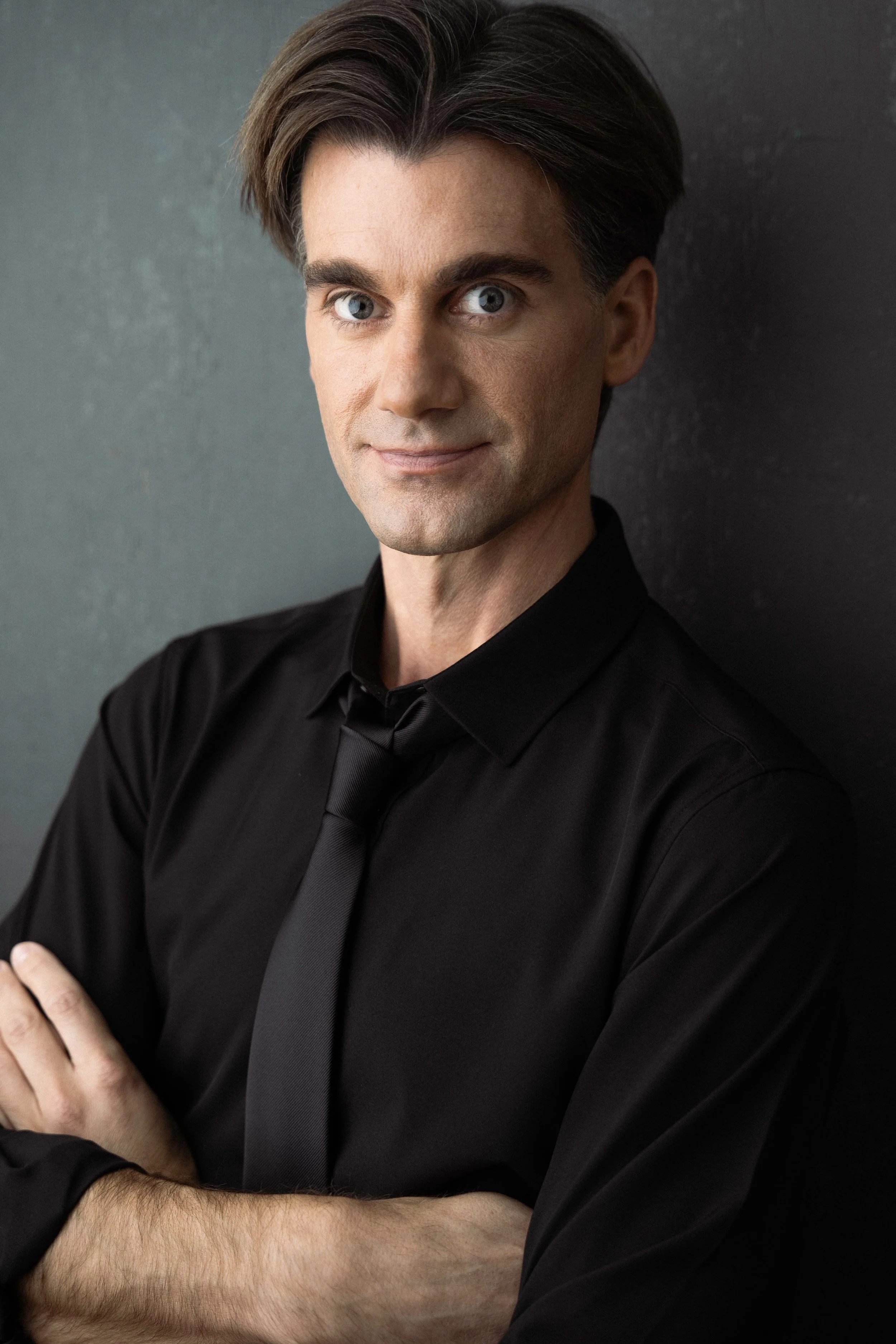A man with short-styled dark hair, blue eyes, and light skin, wearing a black shirt and black tie, standing against a dark gray background. He has a neutral expression with a slight smile, arms crossed, and looking directly at the camera.