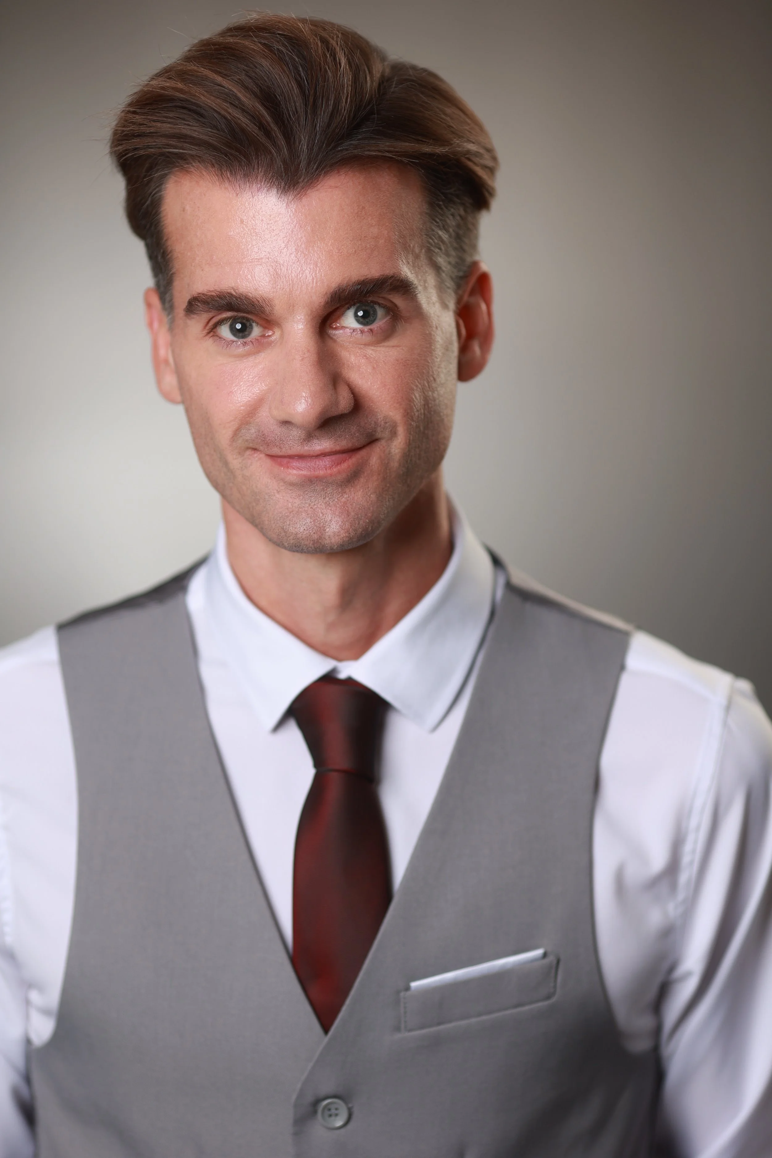 Close-up of a man with brown hair and blue eyes, wearing a white shirt, gray vest, and maroon tie, smiling slightly.