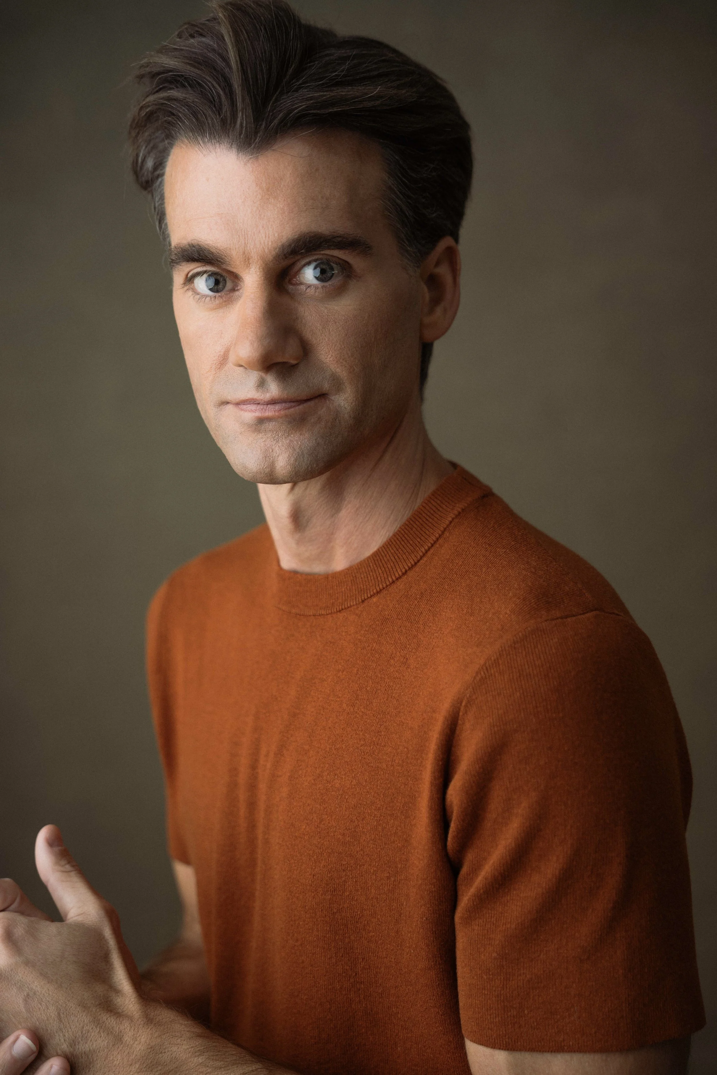 A man with gray hair and blue eyes wearing an orange shirt, looking directly at the camera against a plain background.