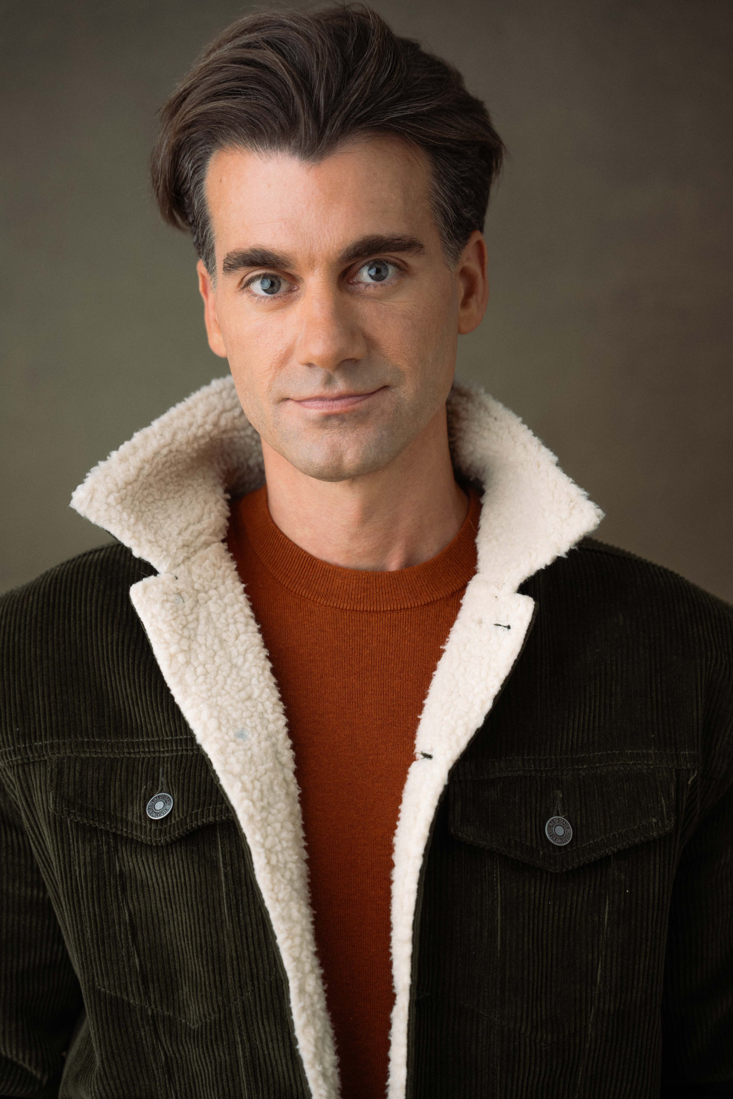 A man with dark hair, blue eyes, wearing a brown shirt and a black corduroy jacket with a cream-colored fleece lining.