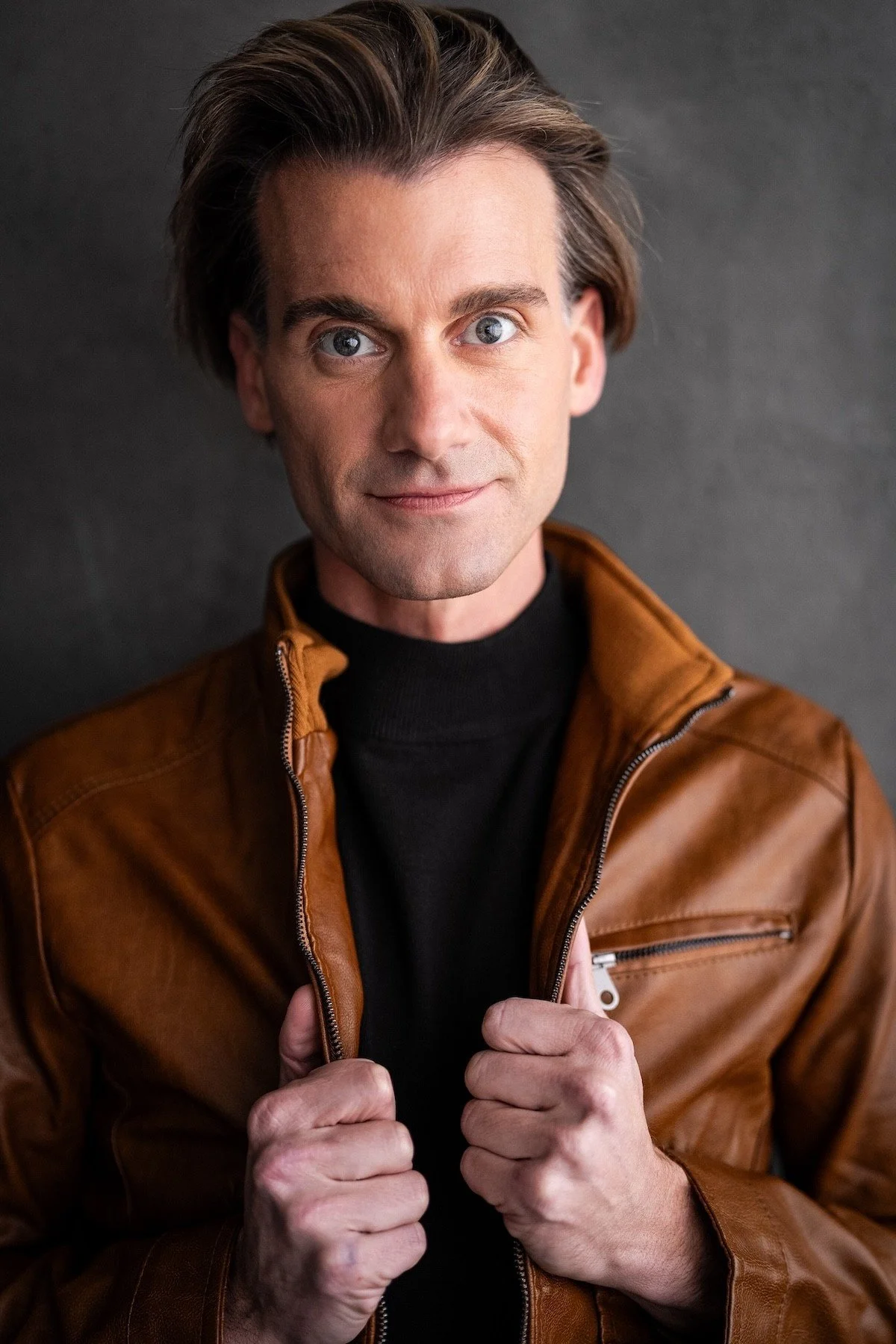A man with light skin and blue eyes wearing a brown leather jacket and a black turtleneck, holding the collar of his jacket with both hands, against a dark background.