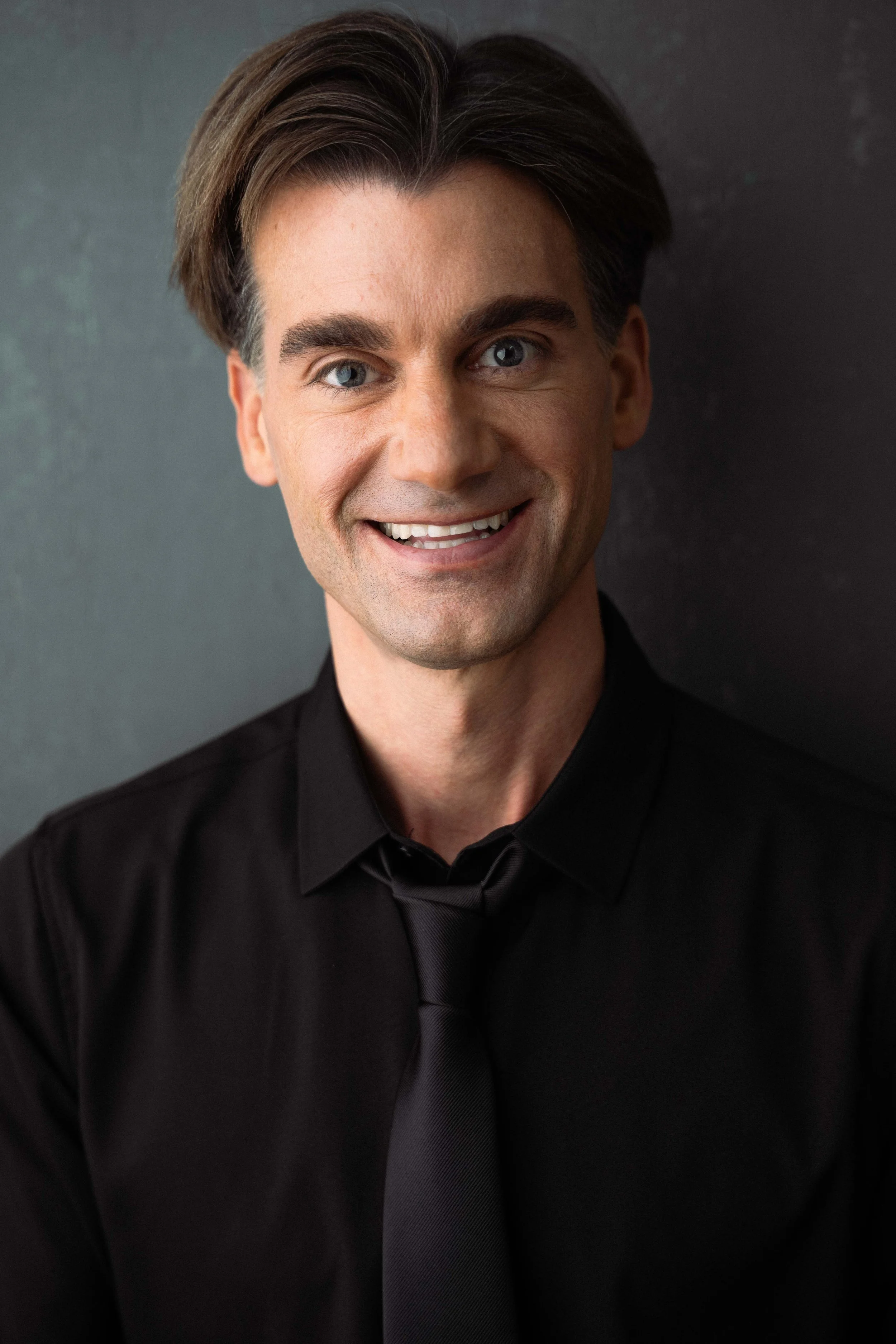 Close-up portrait of a man with brown hair, light skin, blue eyes, and a big smile, dressed in a black shirt, against a dark background.