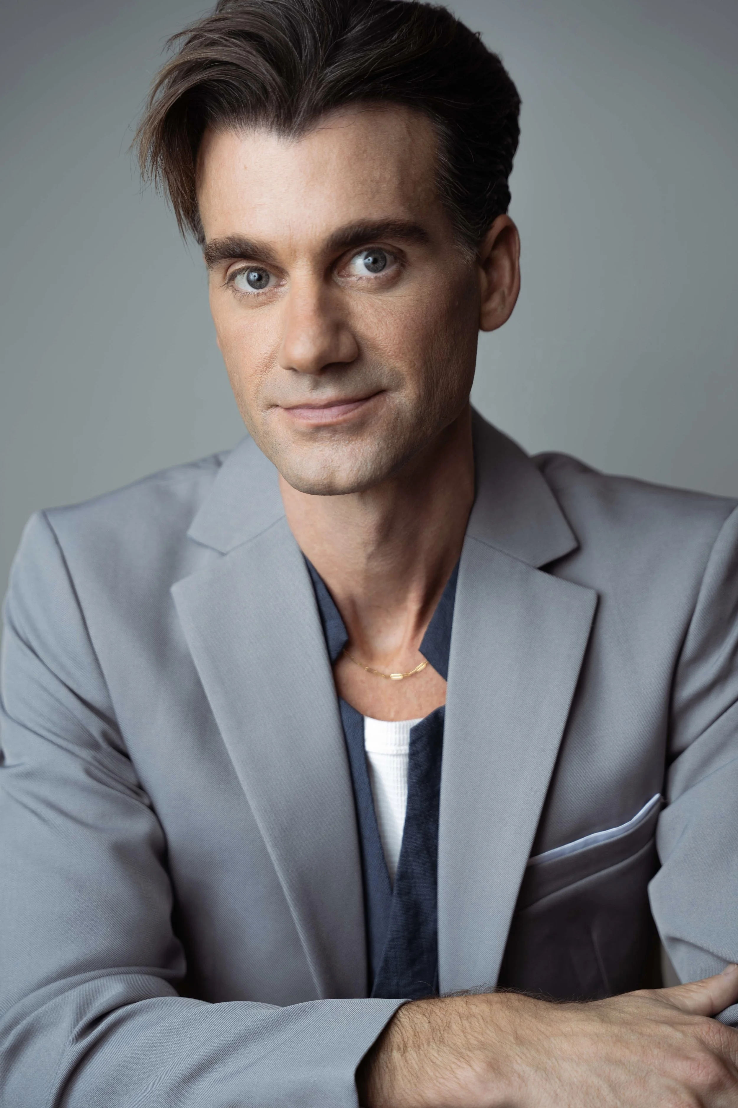 Close-up of a man with short dark hair and blue eyes, wearing a light gray blazer and a delicate gold chain, sitting with his arms crossed against a plain backdrop.