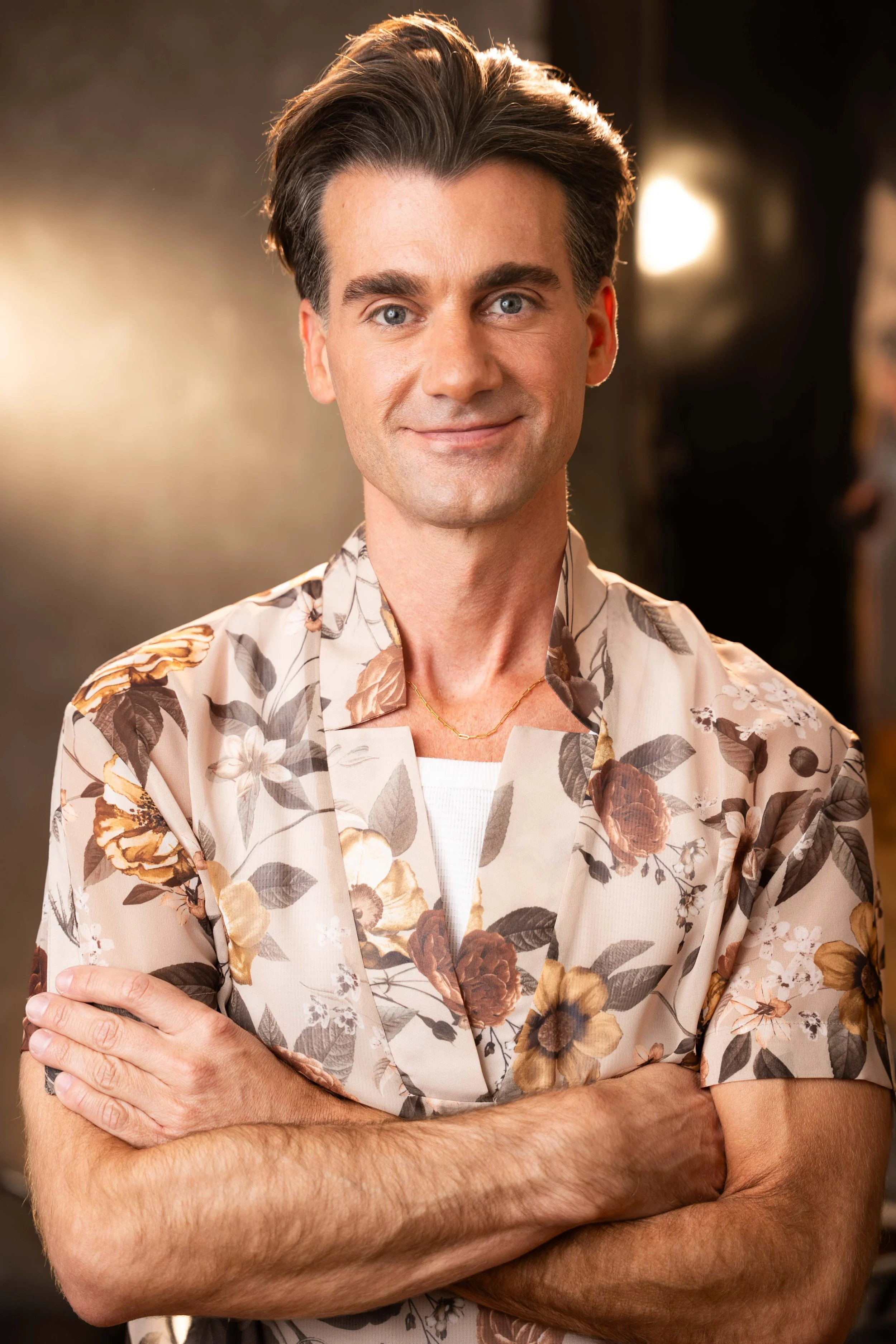 A man with blue eyes and dark hair styled back, wearing a patterned beige floral shirt, smiling slightly, with arms crossed in front of him.