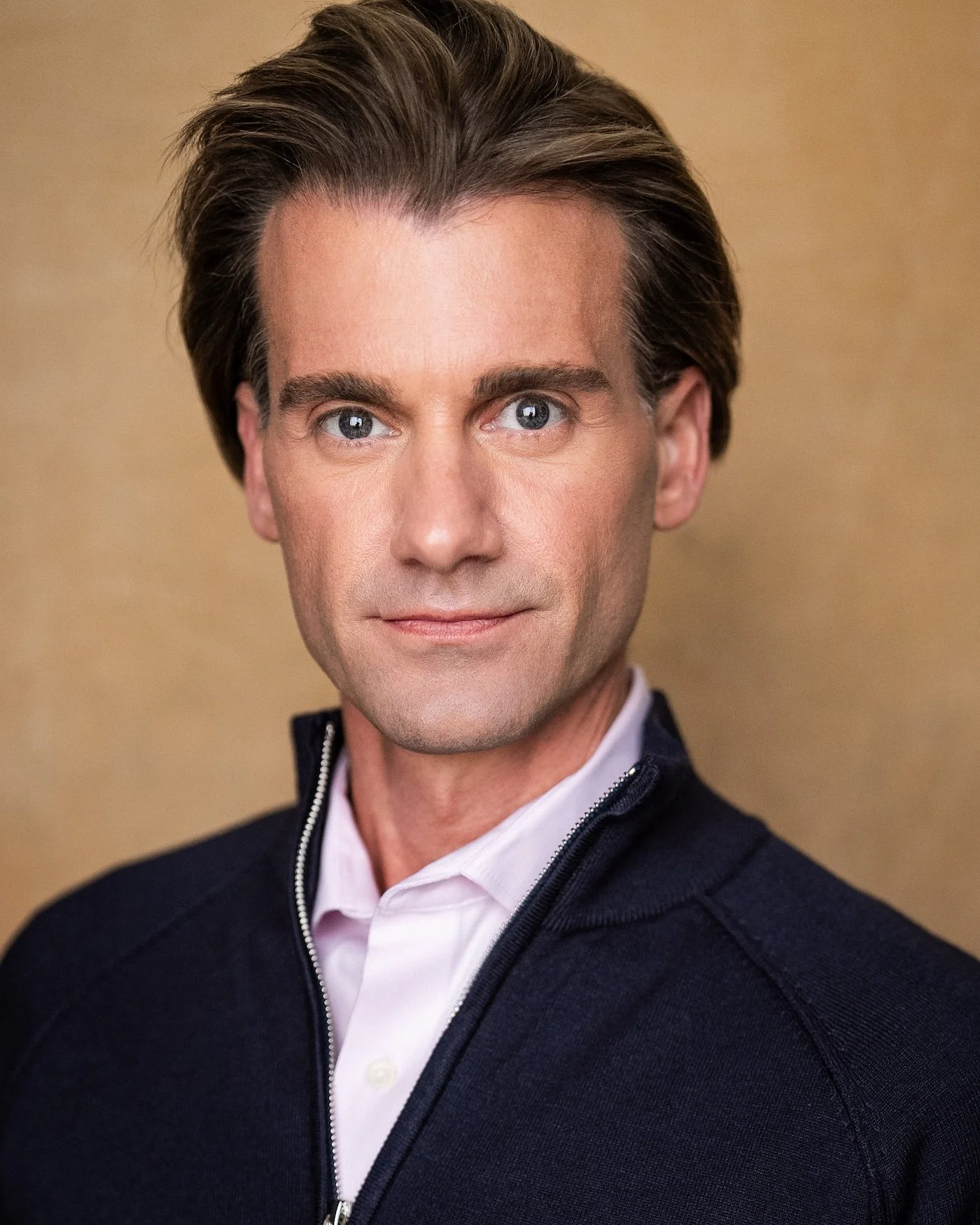 A man with dark hair and blue eyes wearing a white shirt and a dark zip-up jacket, looking directly at the camera, with a neutral background.