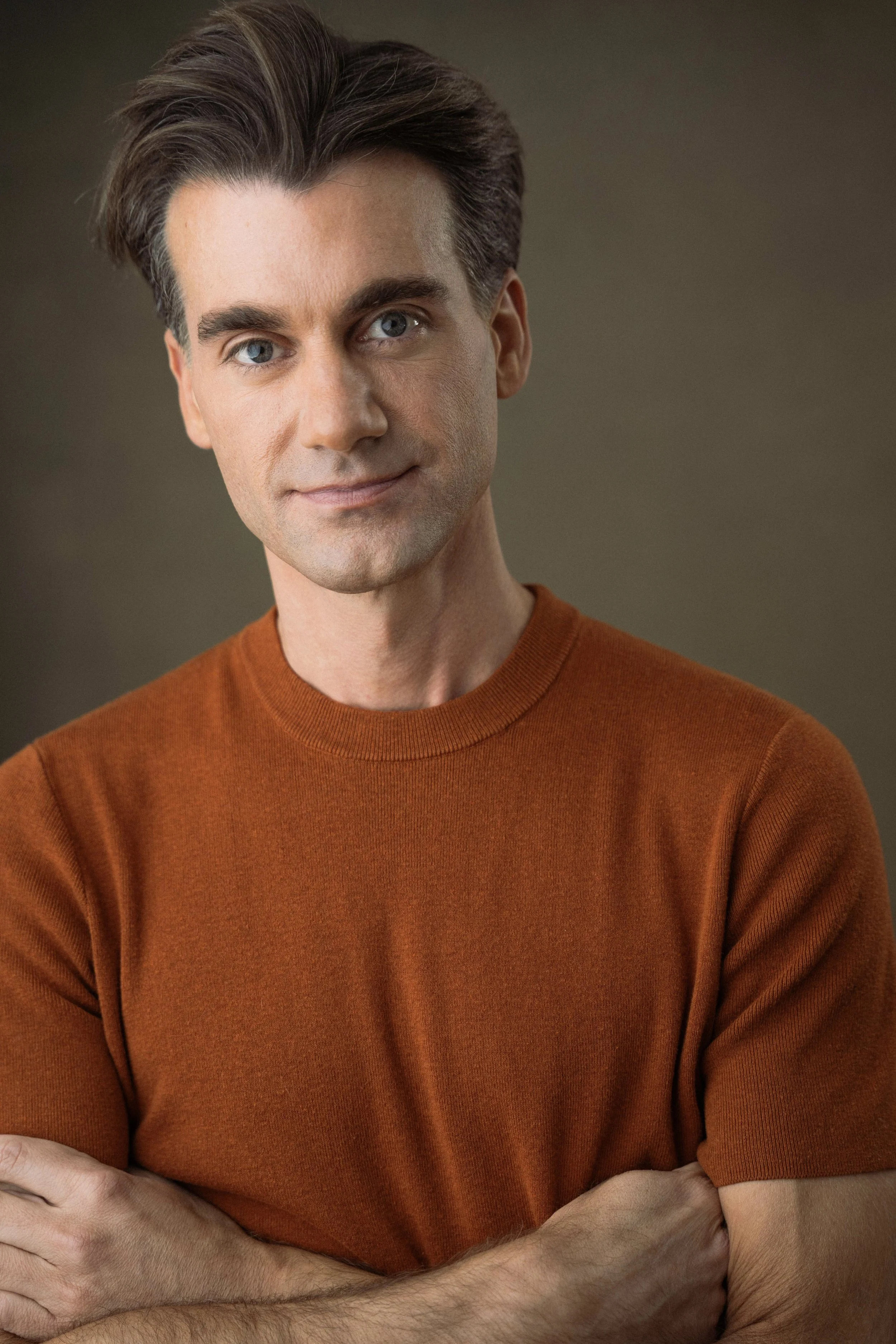 Close-up of a man wearing a burnt orange sweater, with arms crossed, showing only the upper part of his body against a neutral background.