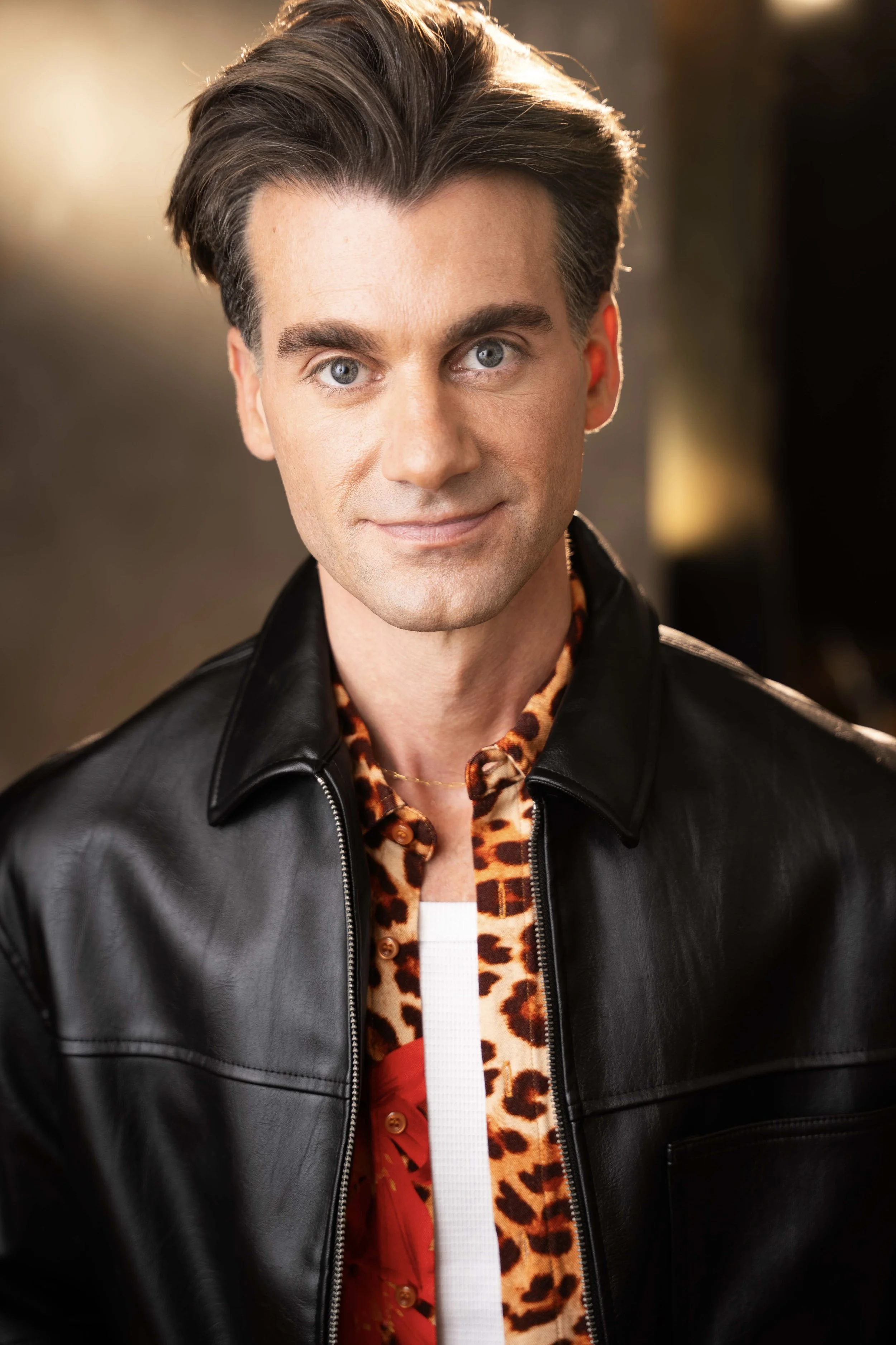 A man with gray hair, blue eyes, and fair skin smiling at the camera. He is wearing a black leather jacket over a leopard print shirt, which is partly visible with a red ribbon or bow. The background is blurred with warm light.